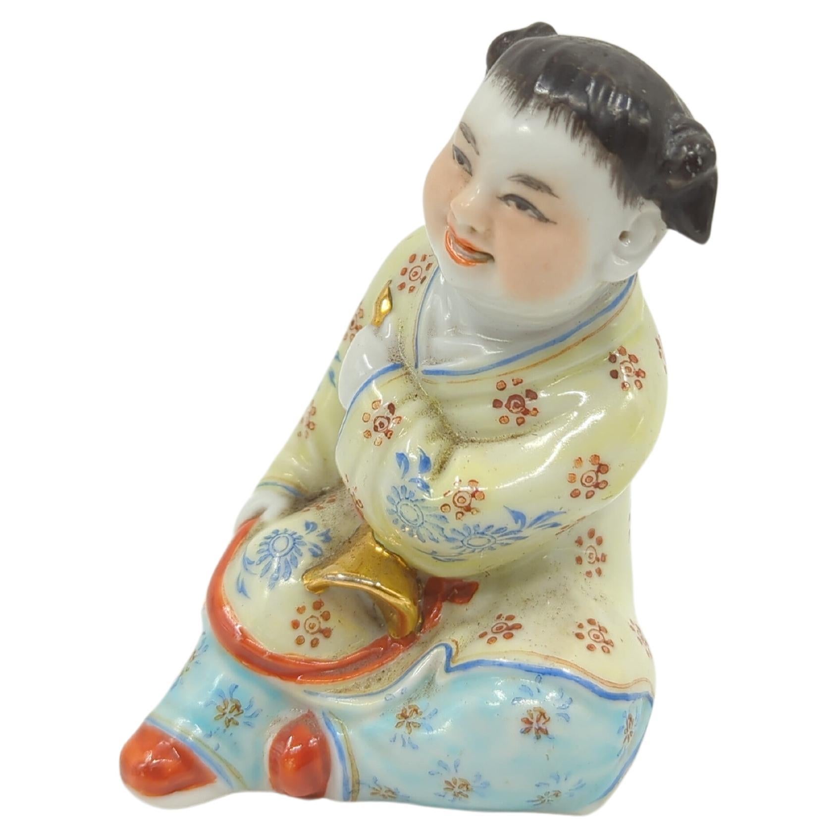 Near antique Chinese famille rose porcelain figurine of a playful, smiling child in a sitting pose, hands holding a golden object likely a instrument, bottom with impressed markings: 