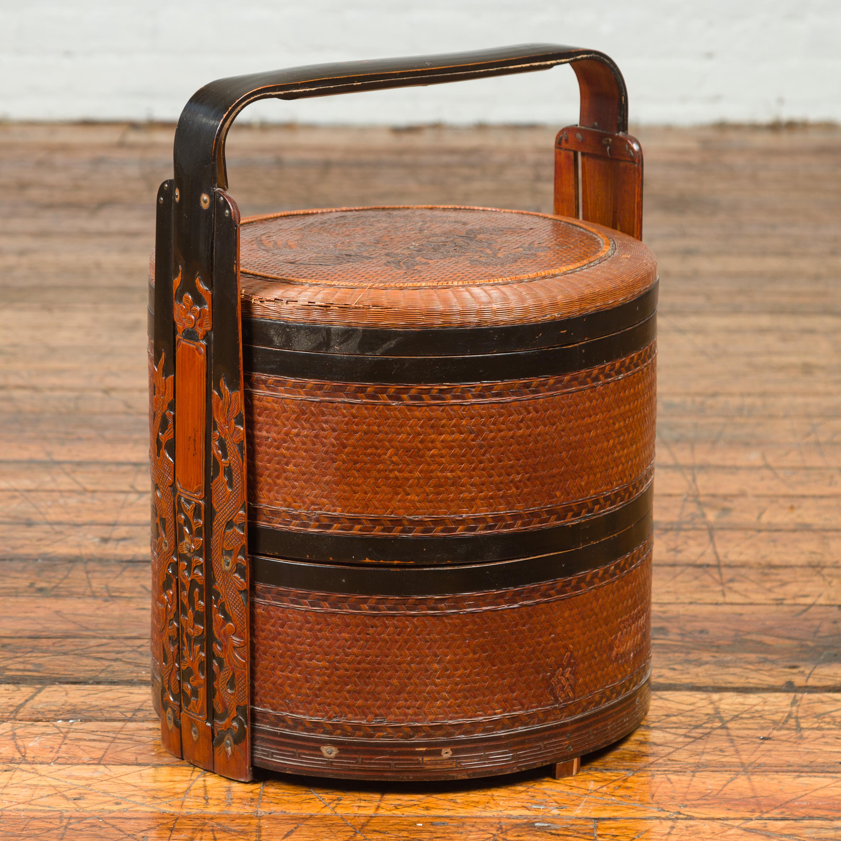 A Chinese vintage rattan and lacquered bamboo two-tiered nested lunch basket from the mid-20th century, with flowers and carved handle. Born in China during the mid-century period, this lunch basket features a circular lid adorned with dark brown