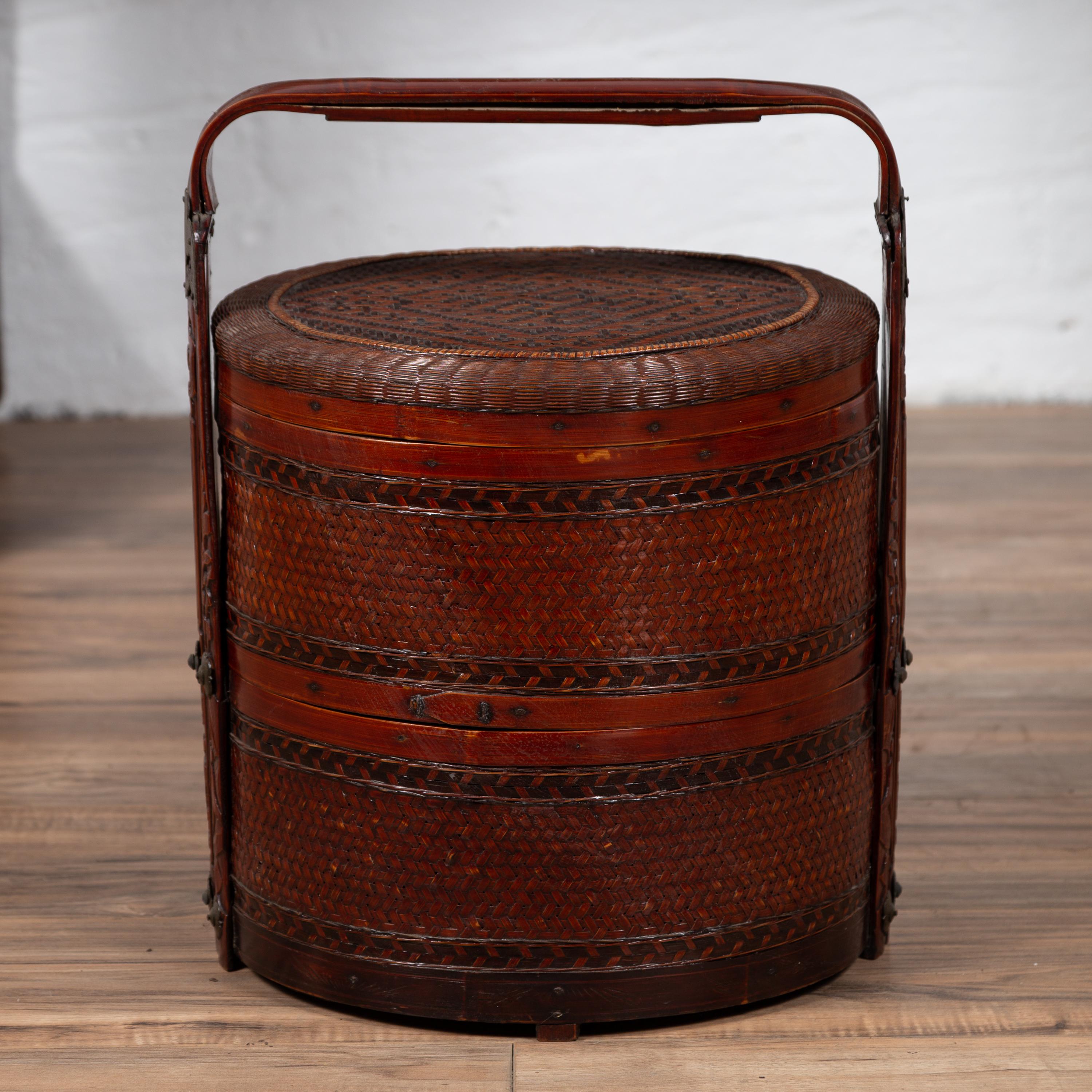 A Chinese vintage rattan two-tiered nested lunch basket from the mid-20th century, with geometrical motifs and handle. Born in China during the midcentury period, this charming lunch basket features a circular lid adorned with woven geometrical