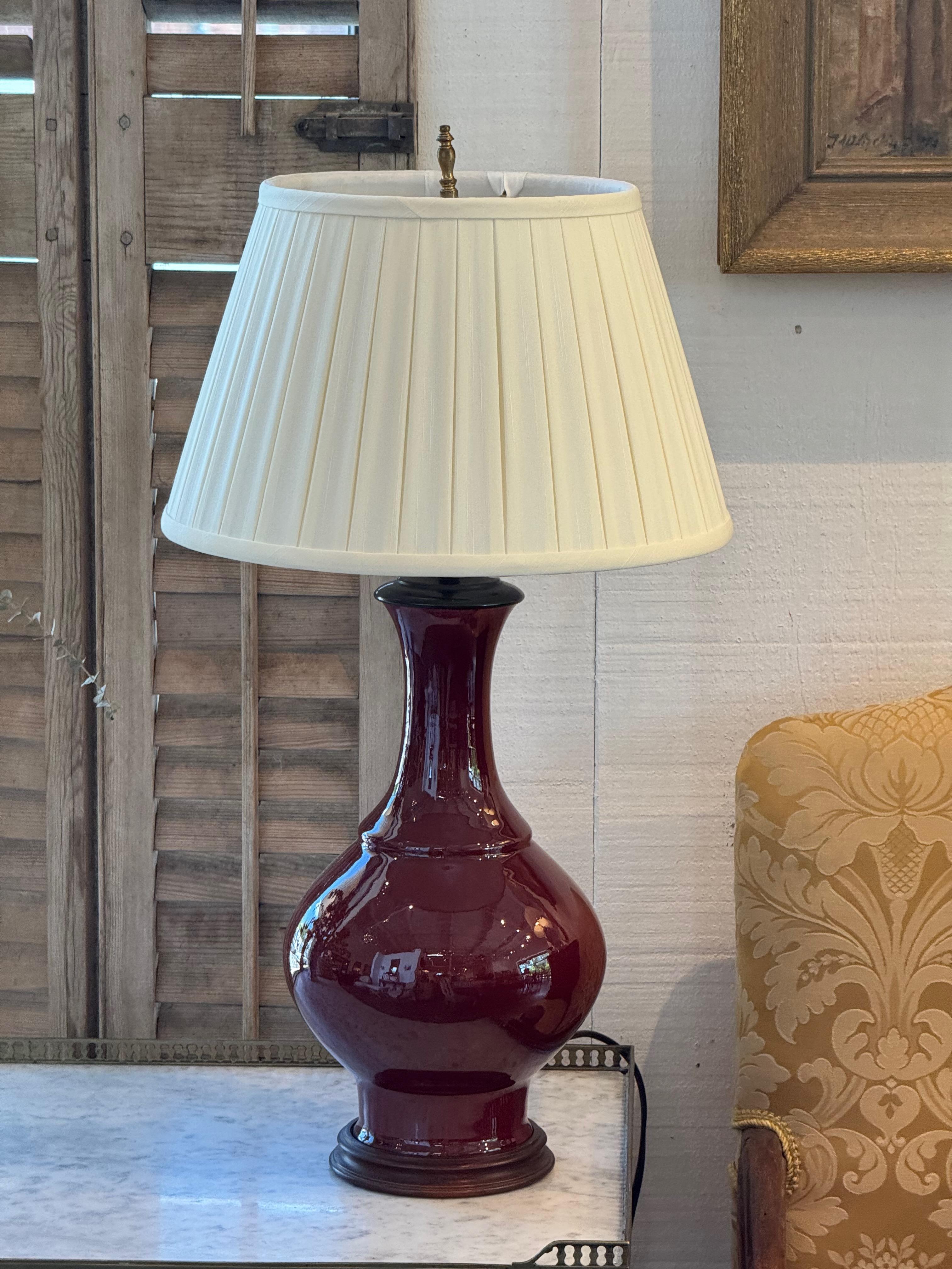 A vintage Chinese table lamp featuring the classic sang-de-boeuf glaze in deep oxblood red. The simple baluster form is paired with a cream pleated shade, blending traditional ceramic craft with understated elegance.