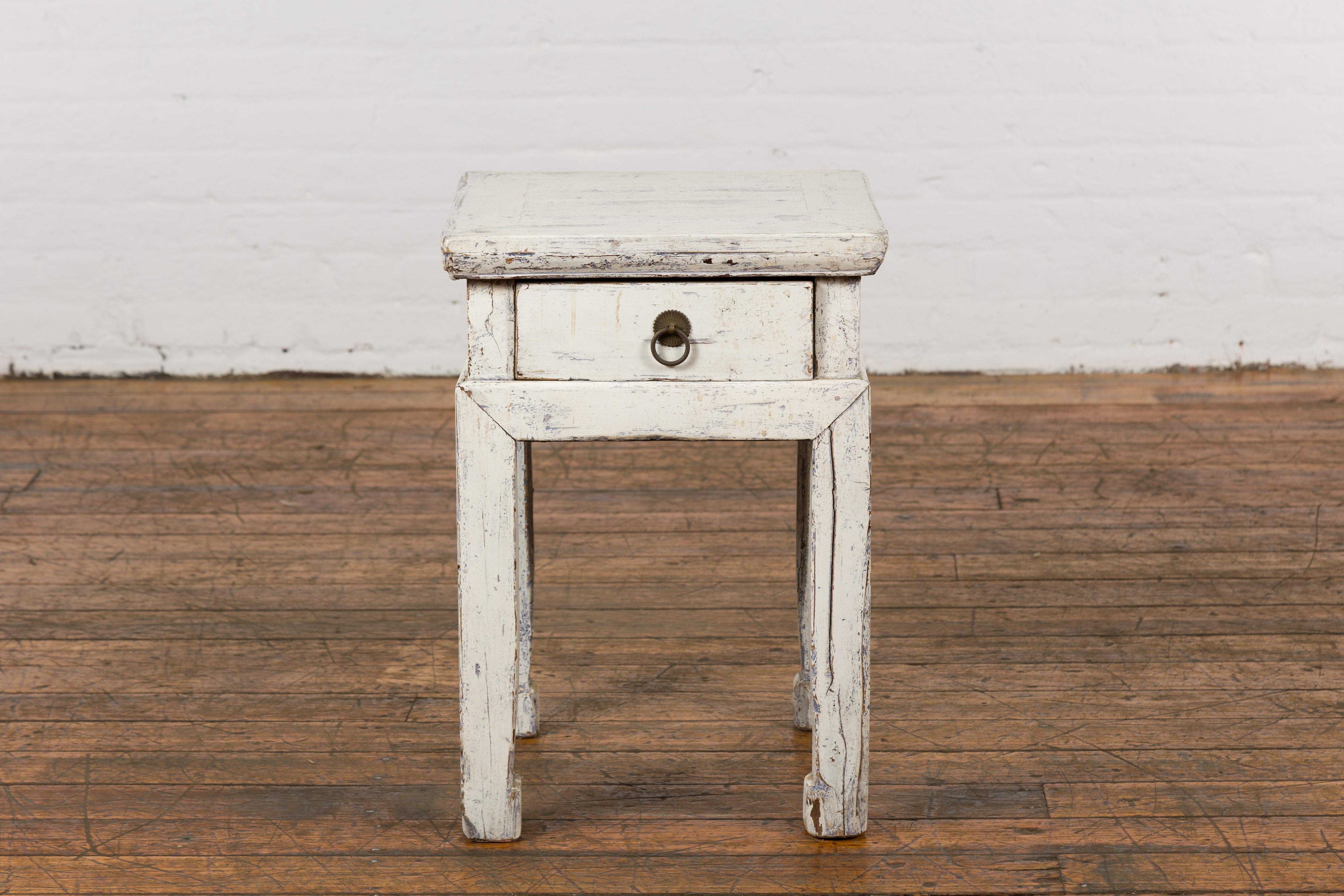 A vintage Chinese side table from the mid 20th century with whitewash finish, single drawer, brass ring pull on floral backplate, horse hoof legs and professionally done custom distressed finish. Imbued with an air of vintage elegance, this