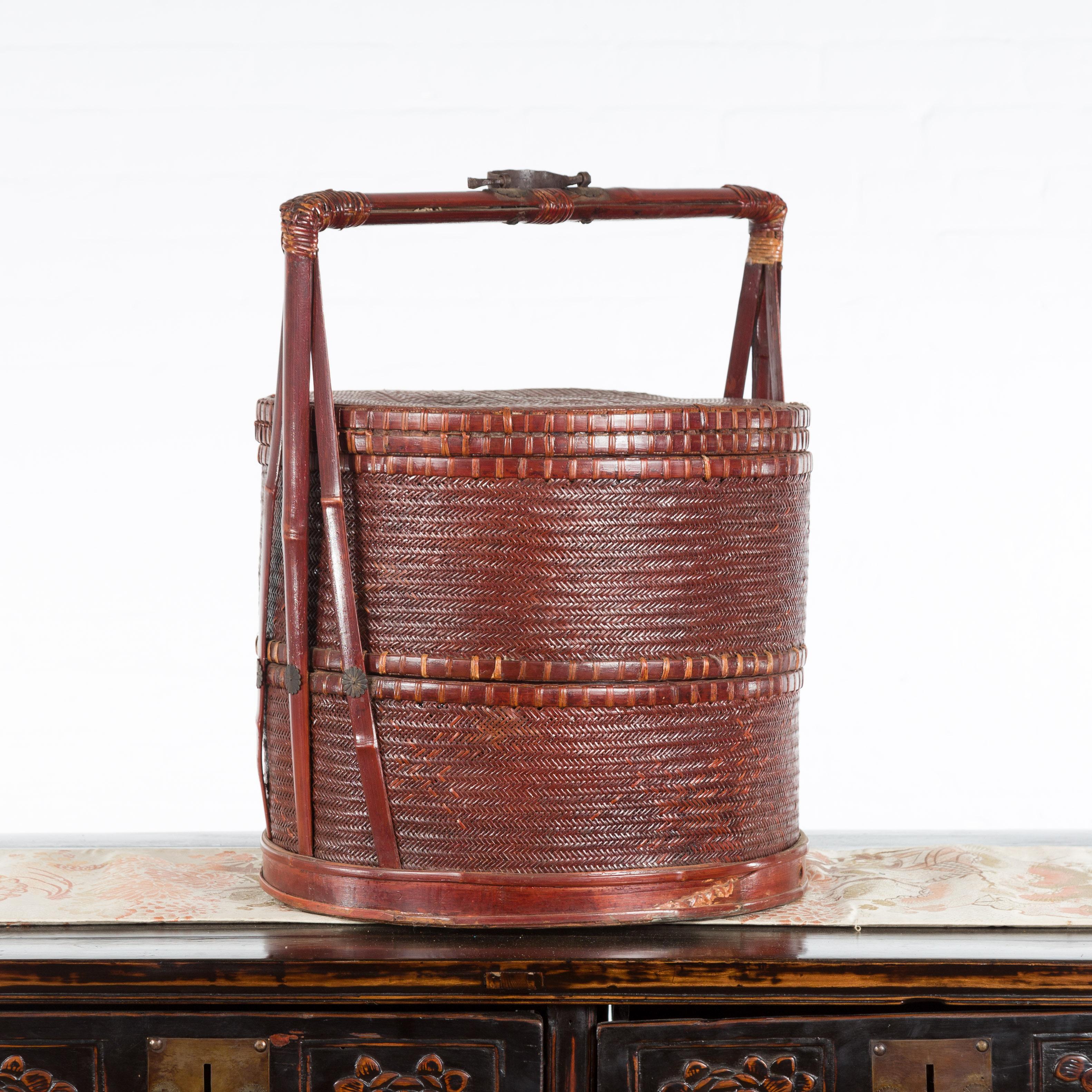 A Chinese vintage woven rattan carrying basket from the mid 20th century, with bamboo handle. Created in China during the midcentury period, this woven rattan carrying basket features a circular lid sitting above two independent sections. The basket