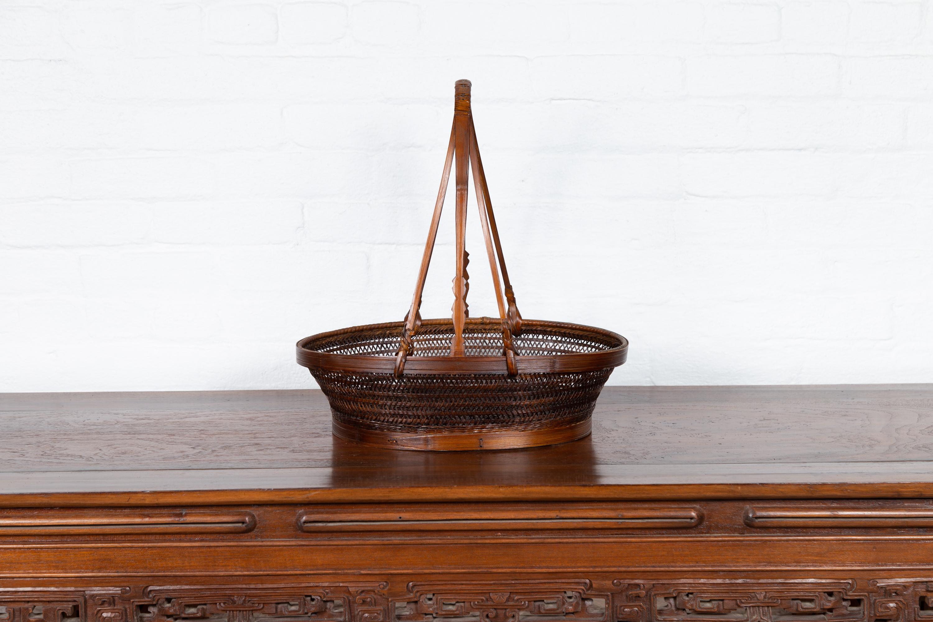 A Chinese vintage woven rattan carrying basket from the mid-20th century, with large handle. Born in China during the midcentury period, this charming carrying basket features an oval body made of woven rattan with wooden frame, topped with a large