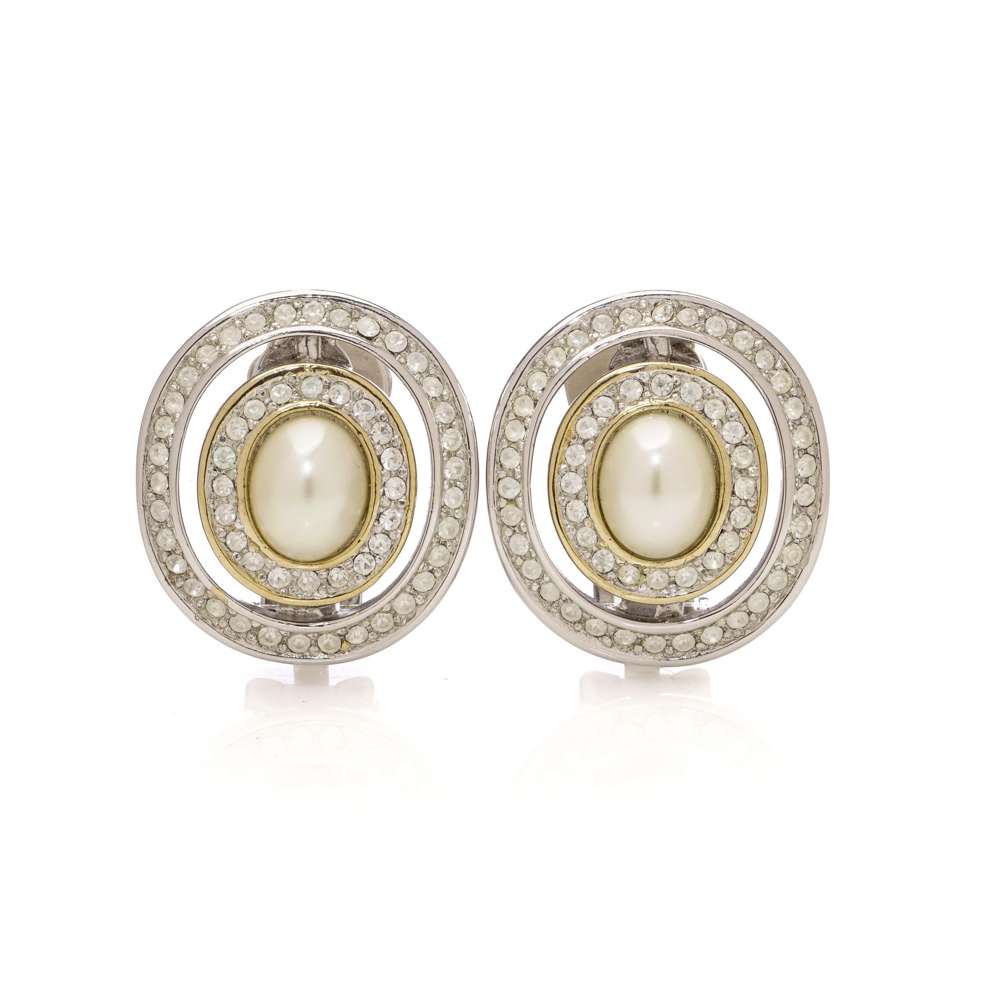 A pair of vintage Christian Dior clip-on earrings, crafted circa 1980s. Each earring is designed with an oval faux pearl cabochon at the centre, framed by a double halo of clear crystals in a silver and gold-tone setting. The openwork circular form