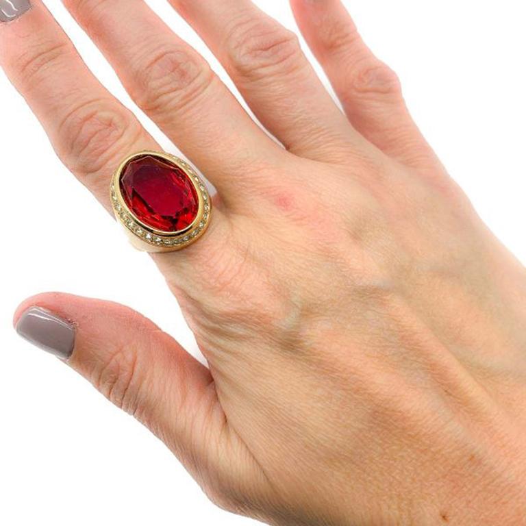 A Vintage Christian Dior Ruby Ring. Featuring a large red faceted oval ruby crystal surrounded by a single row of crystal rhinestones in a gold plated setting. Approx. UK ring size N with an adjustable style band allowing a small size alteration. We