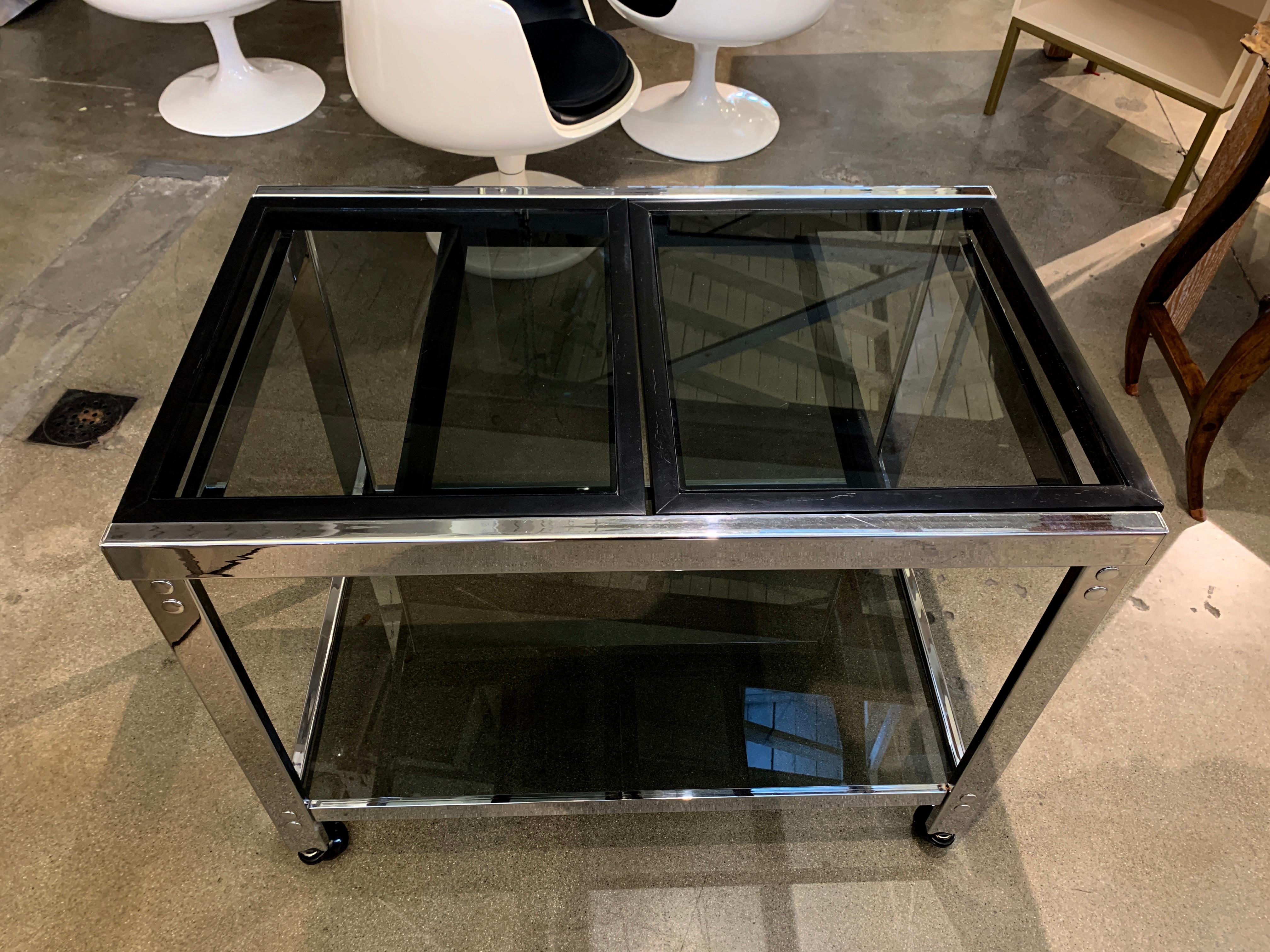 A vintage chrome bar on castors with smoked glass and a slide out top. The top two wood frame tops slide out to make the bar 53 inches wide. It is 36 inches wide when closed. The wood has been repainted at some point black, and there is age