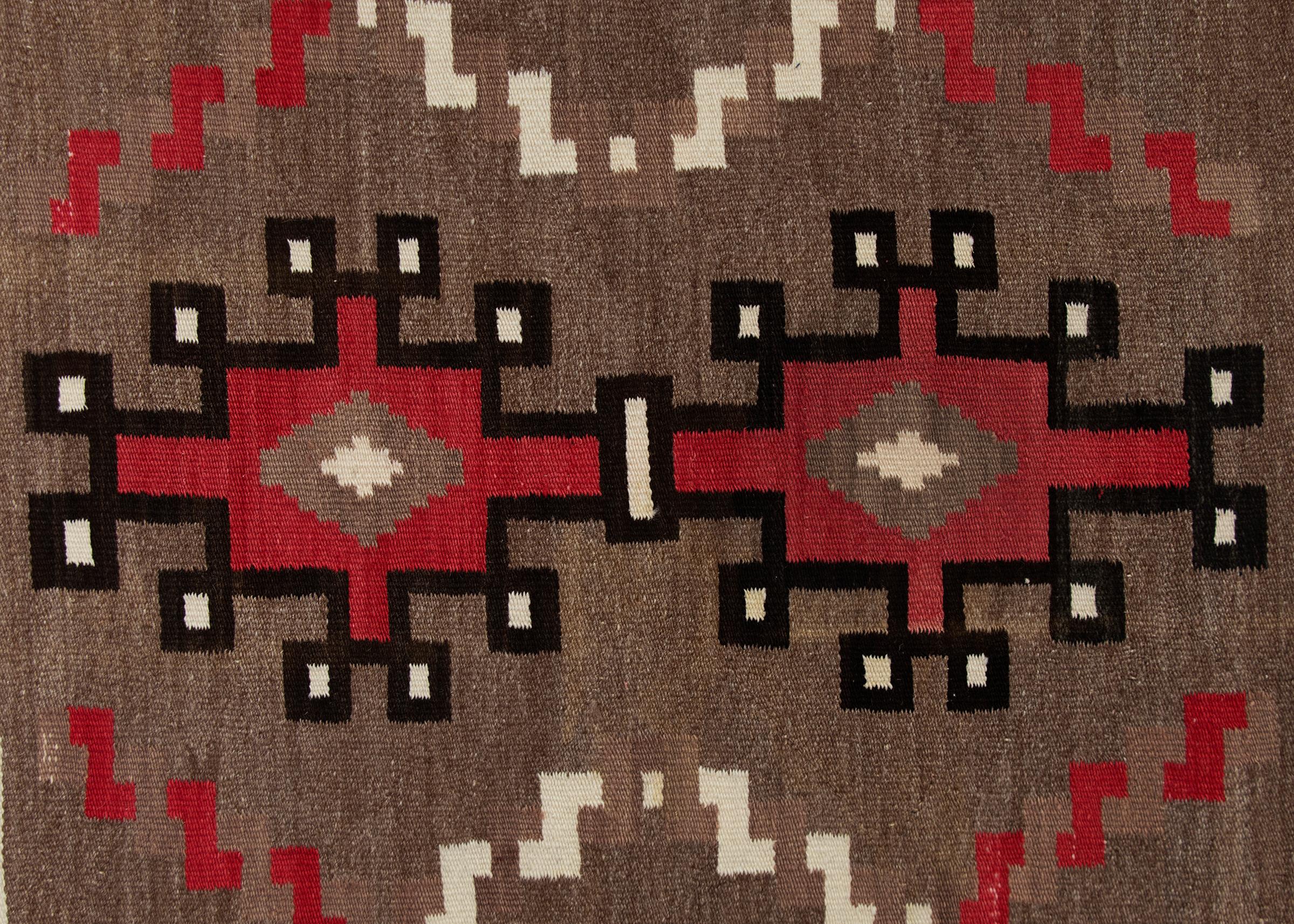 Vintage circa 1940s Navajo rug, handwoven from native hand-spun wool in natural fleece colors of brown/black, ivory and aniline dyed red. Hooked geometric elements are bordered by a tumbling block design. 
The Diné (Navajo) peoples are a Native