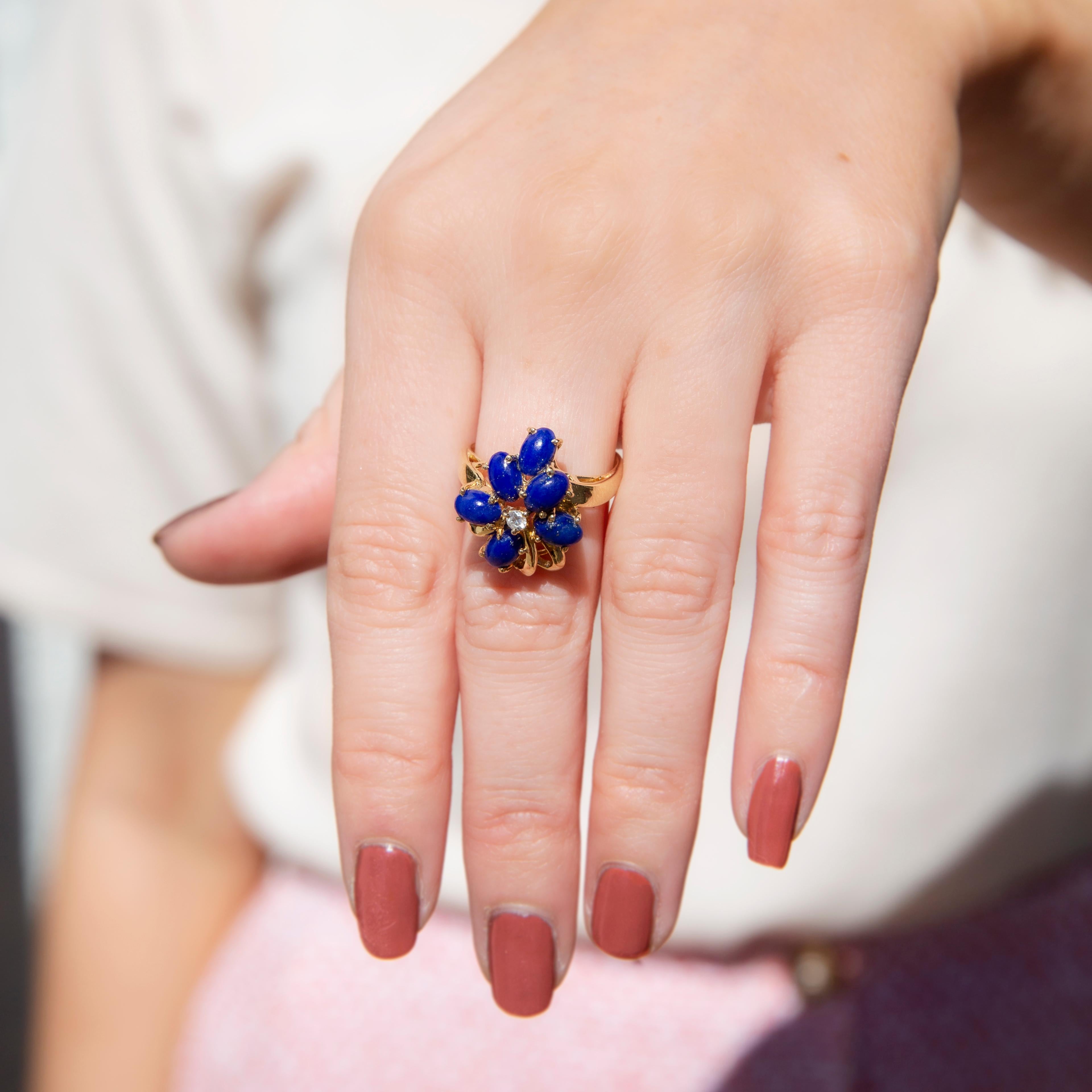 Forged in shimmering 14 carat yellow gold, this decadent vintage crossover ring, circa 1980s, features an asymmetrical cluster of six vivid oval lapis lazuli cabochons with a single round brilliant cut diamond glistening at the centre. This truly