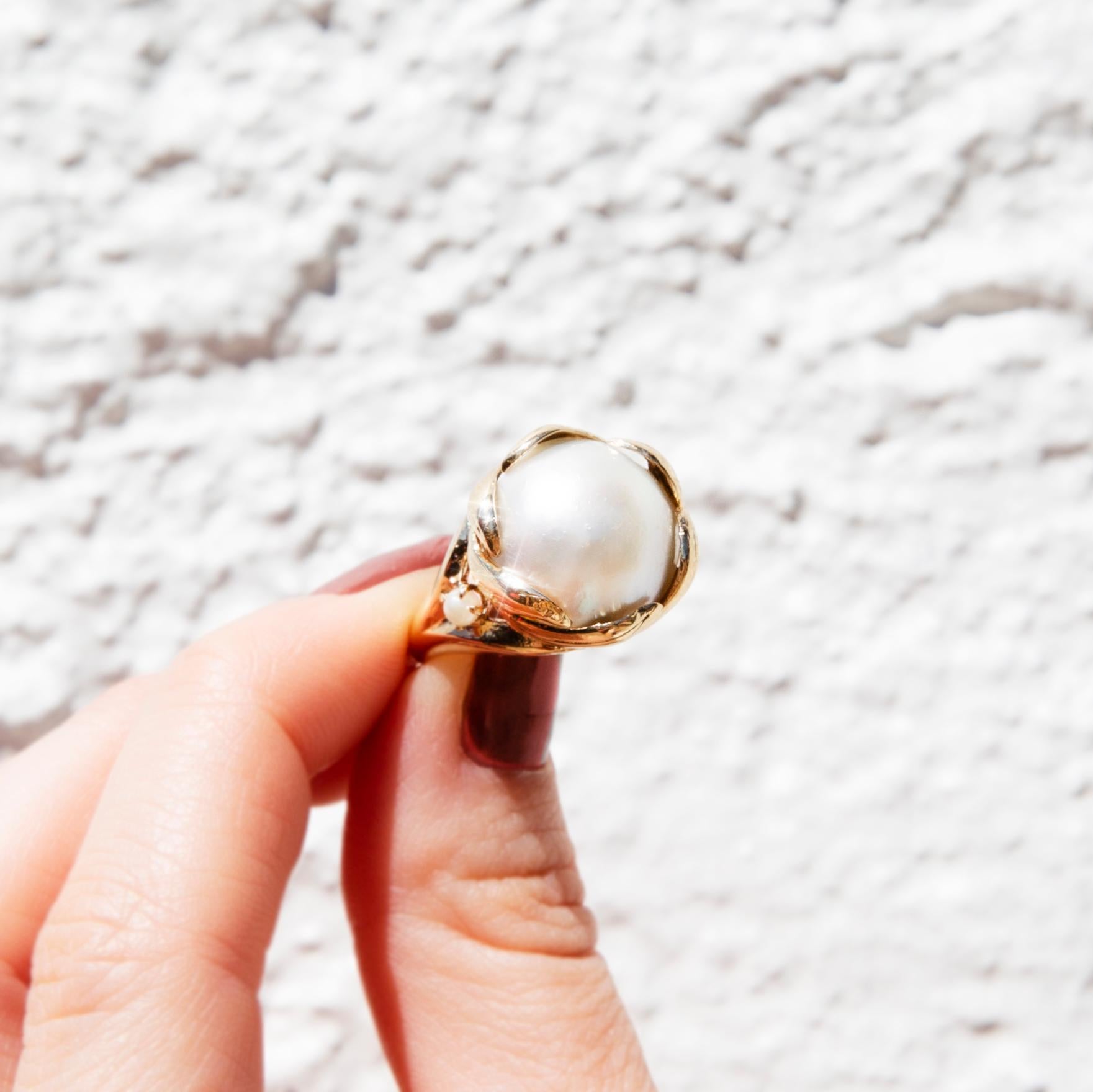 Crafted in 14 carat yellow gold, this darling vintage ring, circa 1980s, features a lustrous mabe pearl on a graceful basket setting, holding two shoulder-set small pearls, that wraps the pearl in elegant leaf-style fixtures. We have named her The