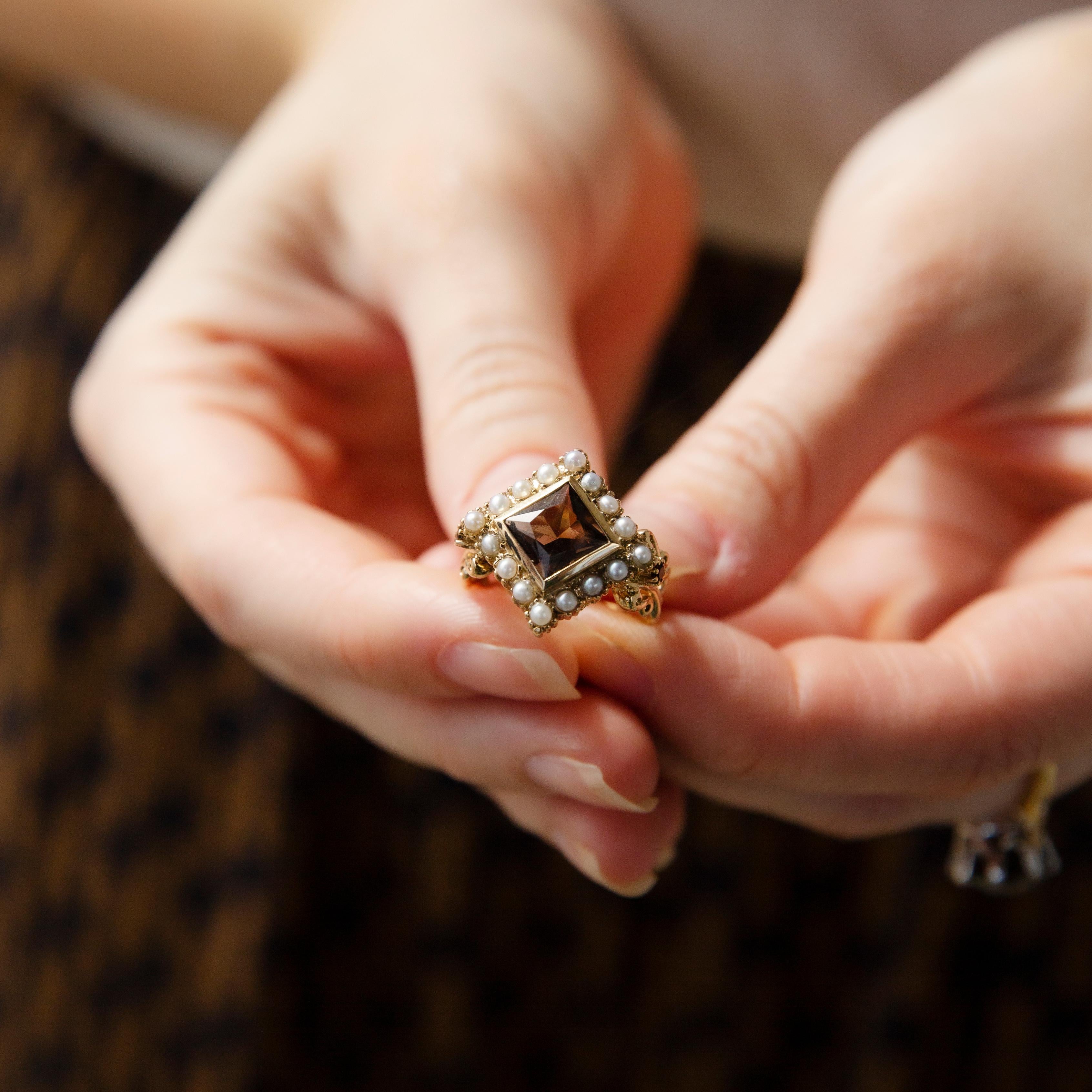 The Nellie Ring, crafted in 9 carat gold, is a charming offset square design that provides it an eccentric twist. Scrolls of ribbon embellished with a single quartz on either side rise to meet a rich bronze smoky quartz framed by lush seed pearls.