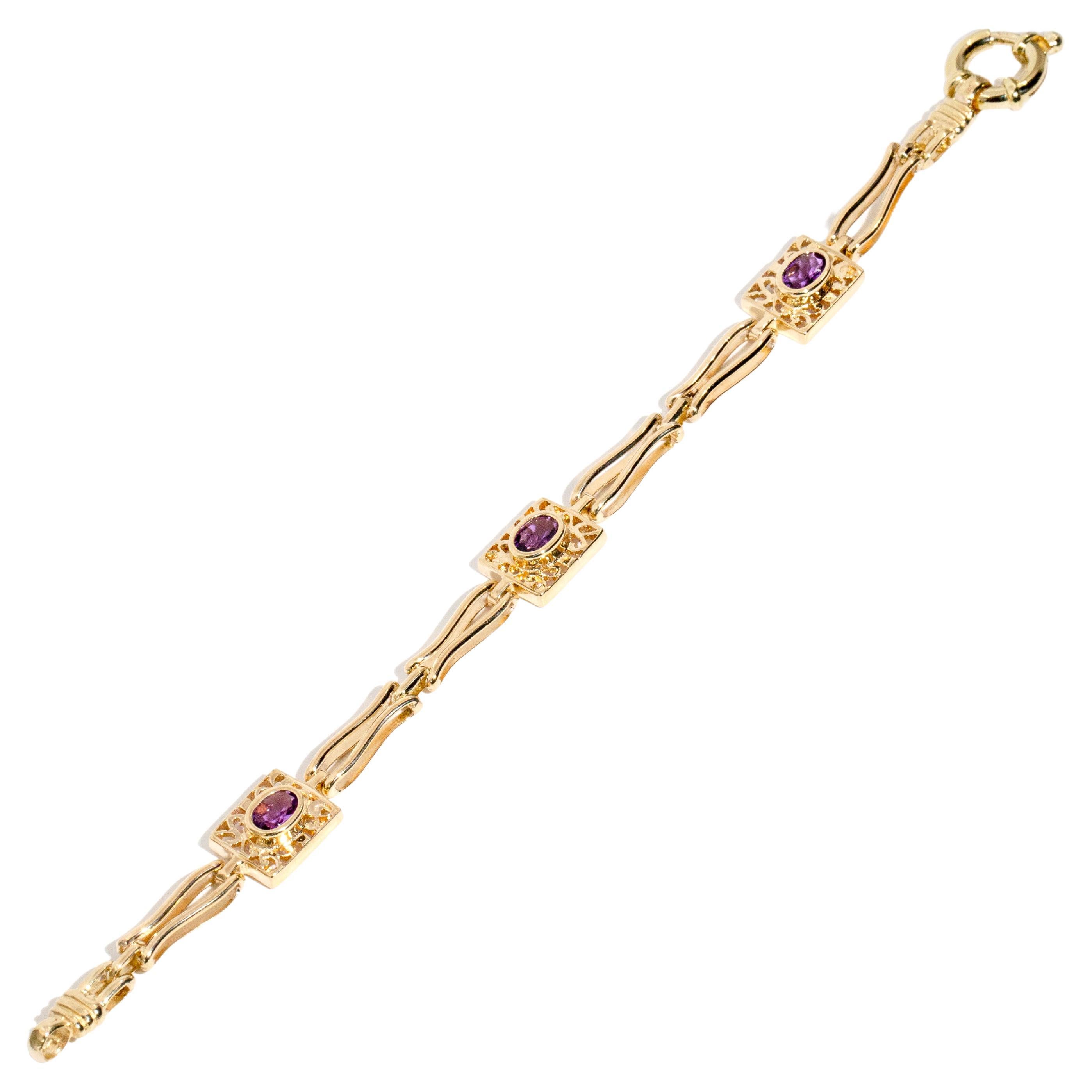Lovingly crafted in 9 carat yellow gold, this charming vintage bracelet, circa 1990s, is a sequence of three filigree settings each holding a raised oval rubover set amethyst and interlinked by pairs of elegant double-bar links. Her name is The