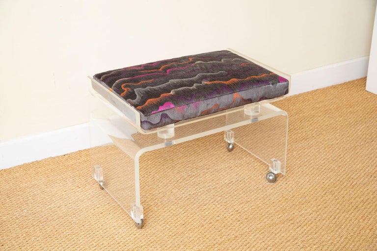 This chic vintage clear lucite vanity bench has 2 tiers under the cushion which has been newly re-upholstered in a chenille colorful abstract fabric with a dark gray background infused with magenta, purple, light gray and some white with accents of