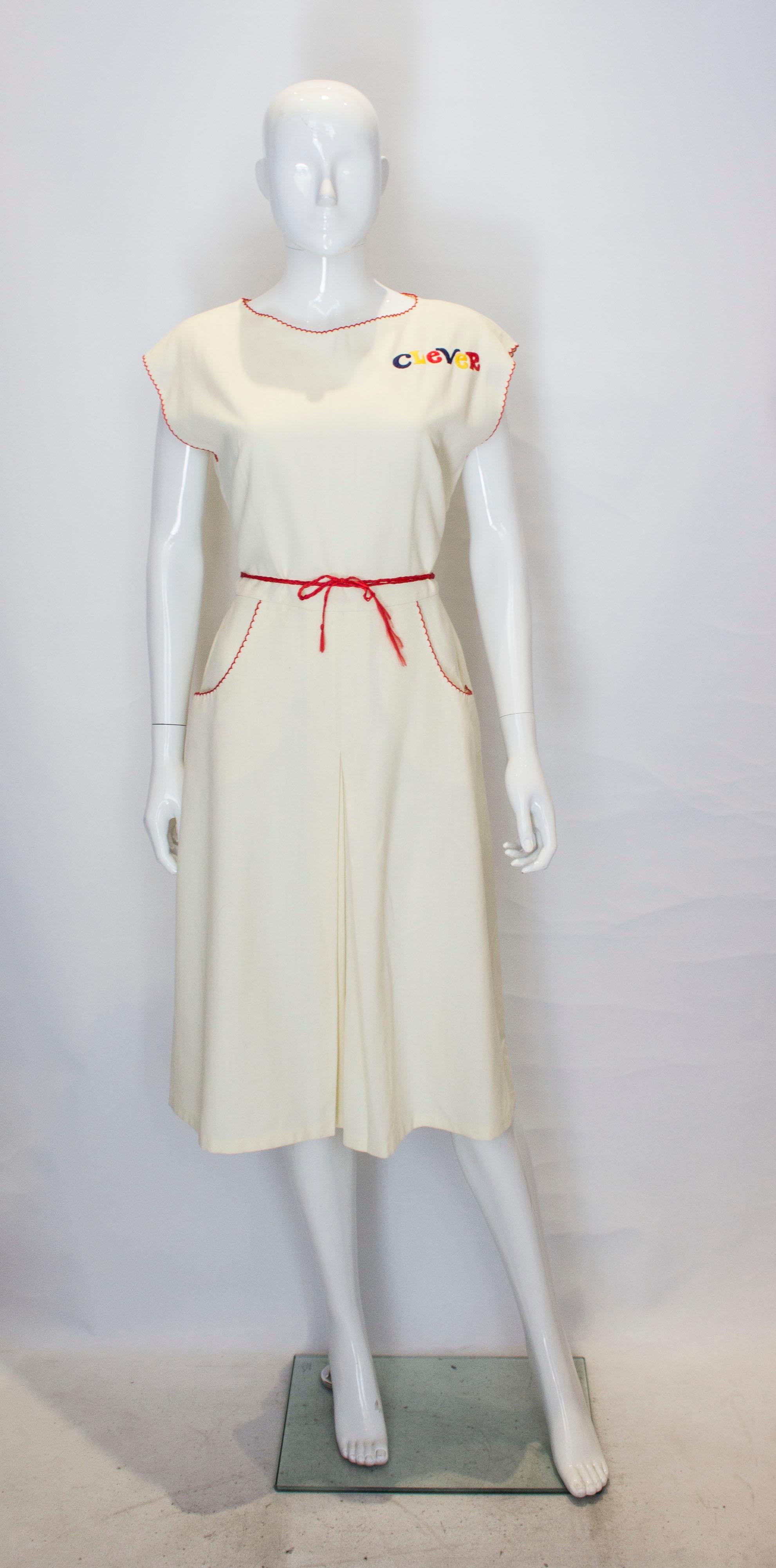 A fun vintage novelty dress with the word 'Clever' embroidered on the top left.  In an ivory fabric edged in red, the dress has a flared skirt with front  central  pleat , two front pockets, central back zip and a red rope belt.