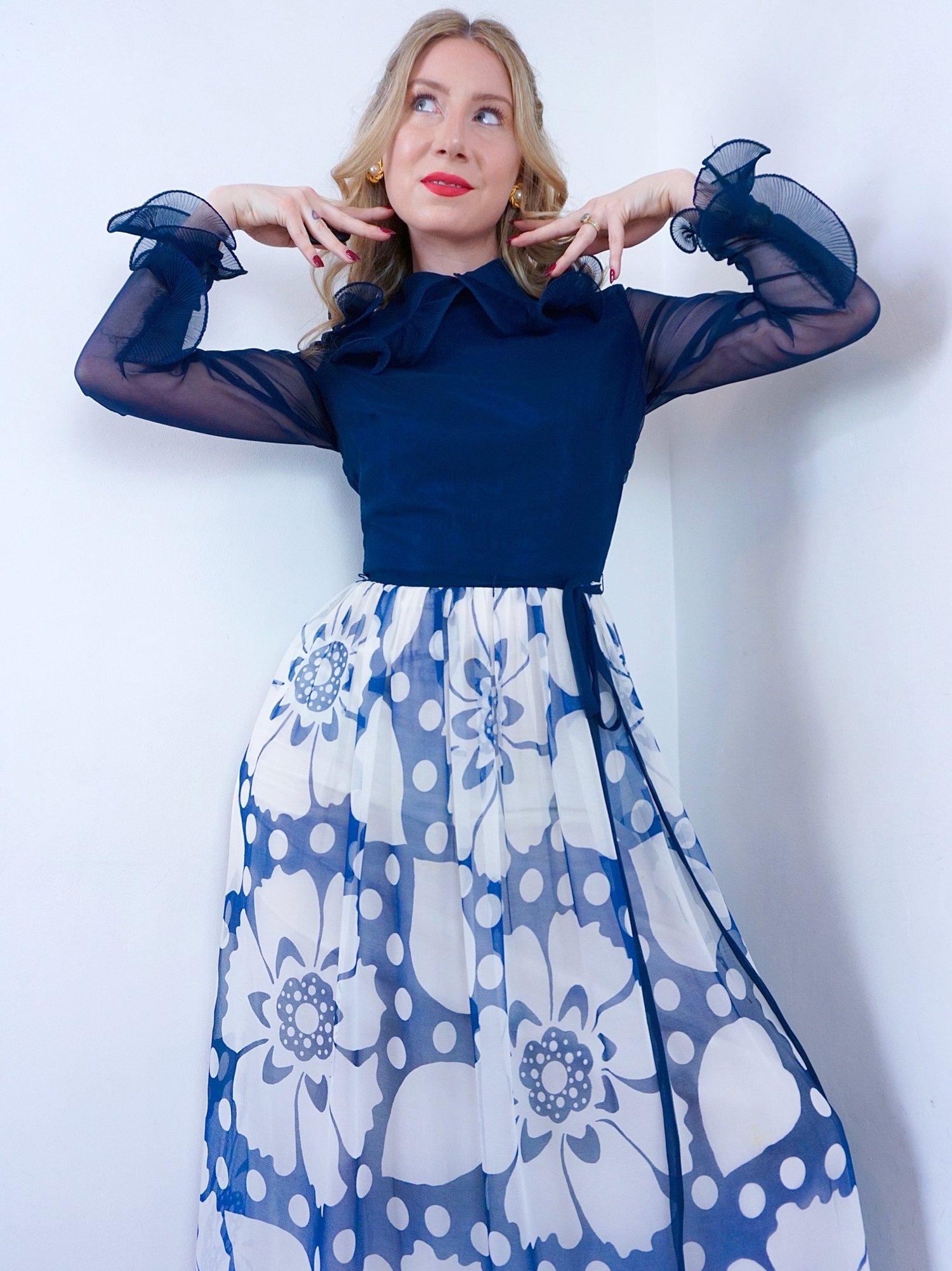 1960s Clive Byrne maxi dress in a navy blue and white floral design. 
She has a dark navy bodice with sheer long sleeves finished with a ruffled cuff that matches her ruffled Peter Pan collar.
A thin tie waist belt cinching in her waist, before