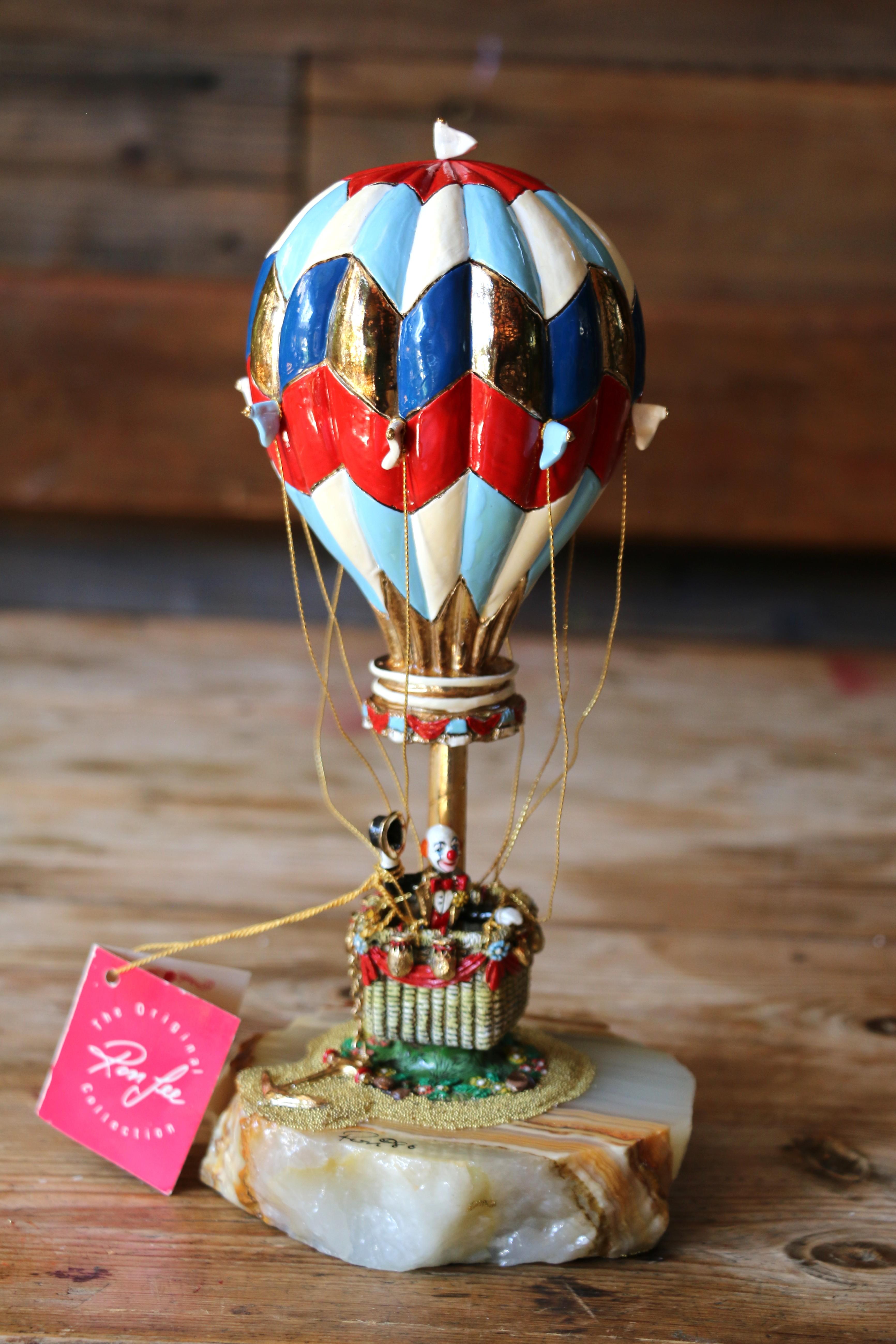 Ti presentiamo questa spettacolare statuetta da collezione che raffigura una mongolfiera colorata con una statuetta di clown nel suo cesto, disegnata da Ron Lee, originale del 1986. Questa meravigliosa scultura è dipinta a mano con colori vivaci