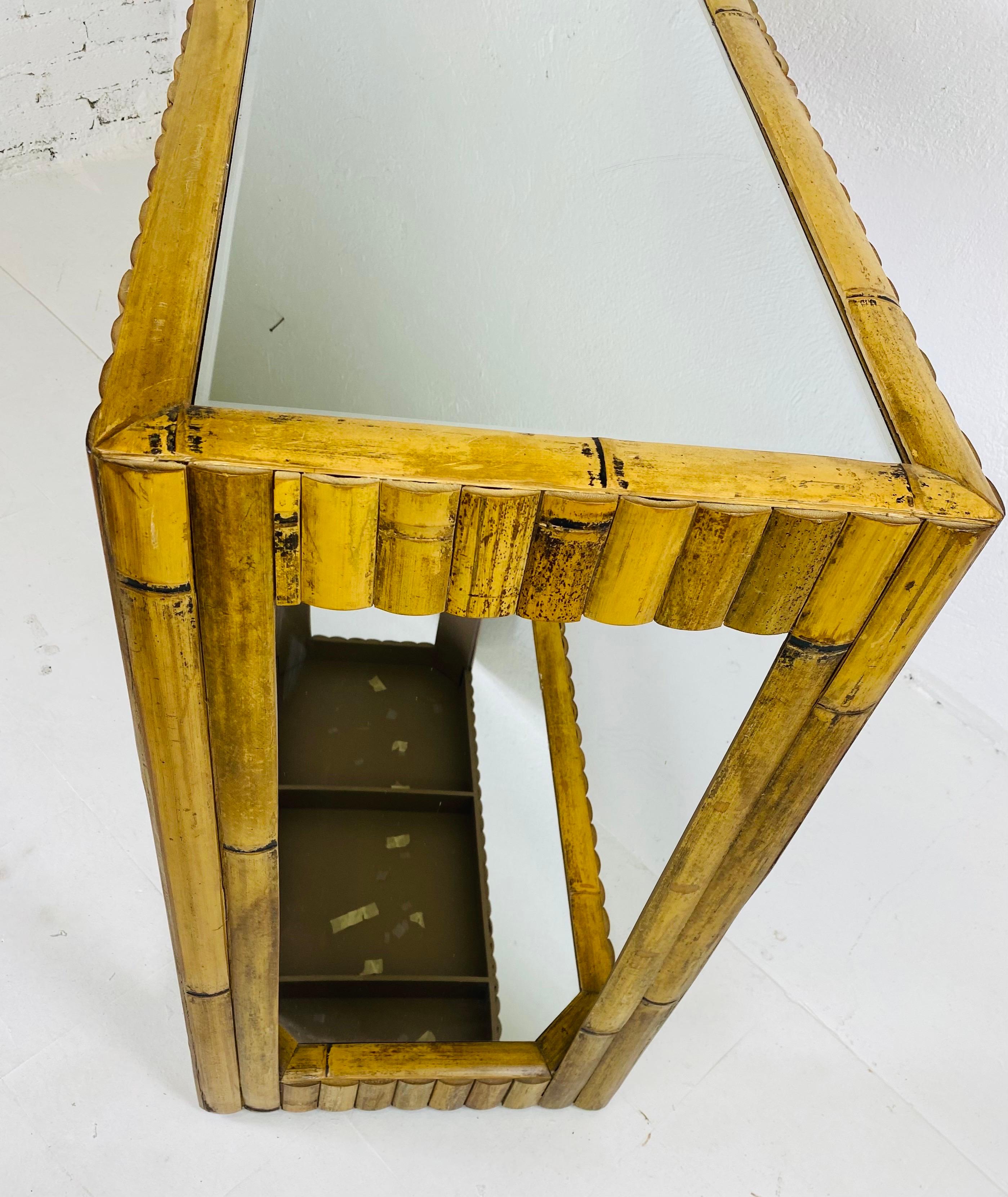 This is a vintage mirrored coastal bamboo console. This console has a mirror top with a single mirrored shelf at the bottom. This console construction has wide halved bamboo planks attached to a wooden frame. This console is great for behind the