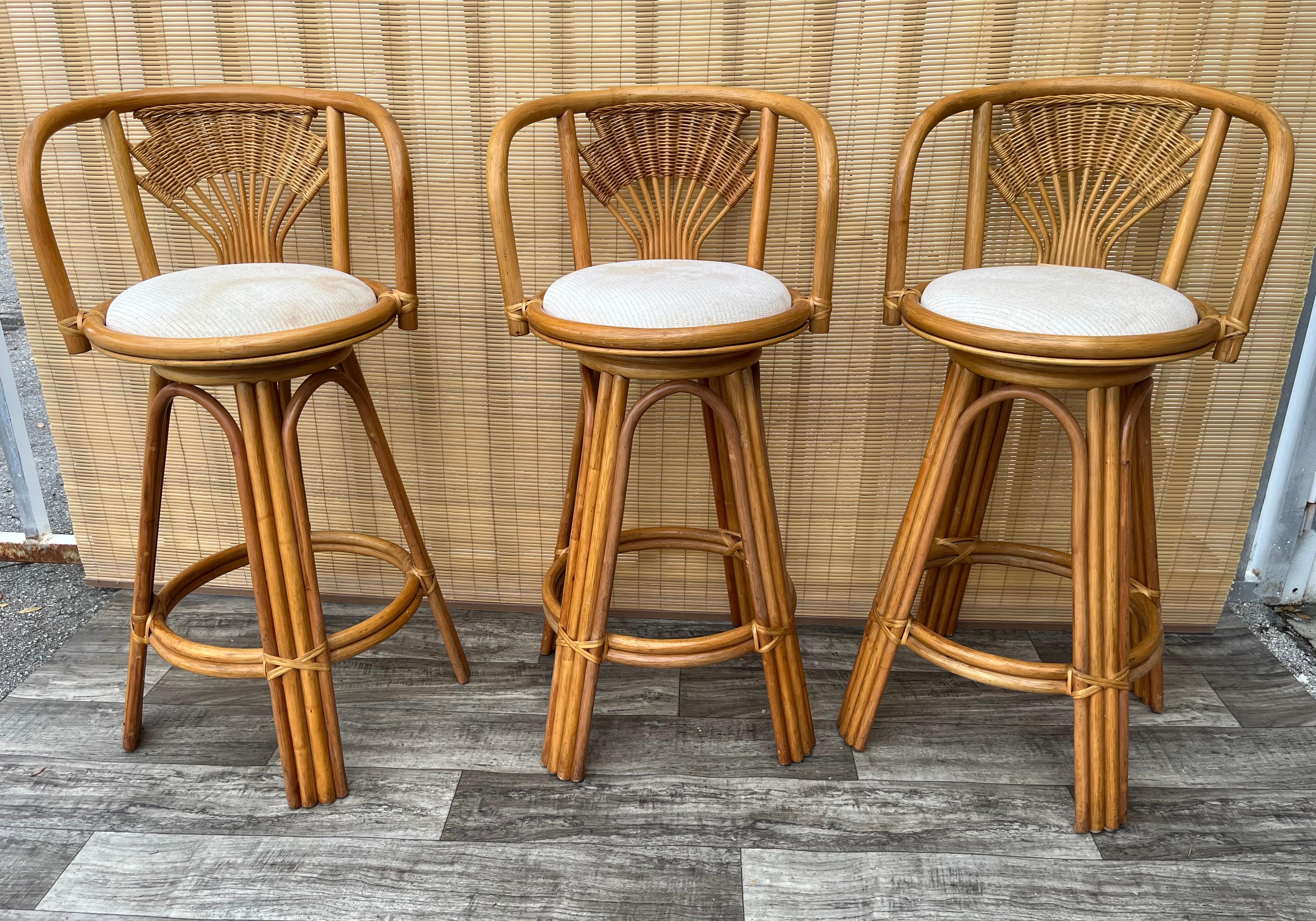 A set of Three Vintage Coastal Style Wicker Rattan Swivel Bar Stools. circa 1980s
Feature a Rattan frame, cane detailing on backrests, and round seats with a the original oatmeal colored fabric seats. 
In good original condition with signs of wear