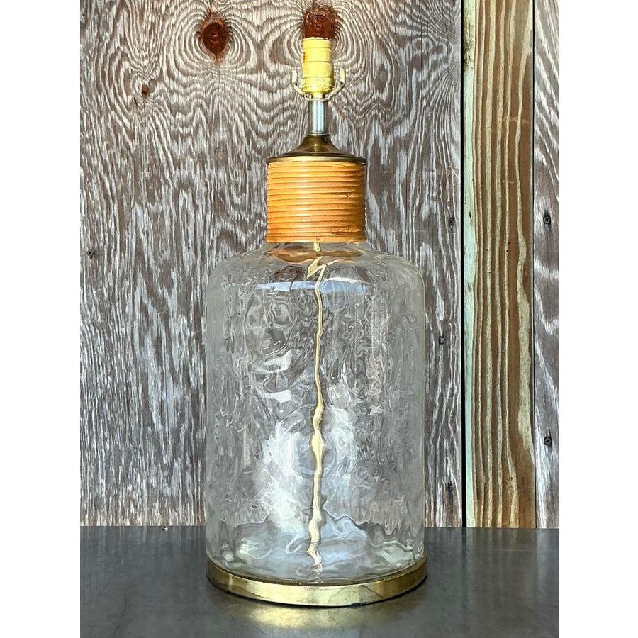 Cette lampe cruche en verre vintage unique présente une élégante base en verre clair avec des motifs texturés subtils qui reflètent magnifiquement la lumière. Surmontée d'un laiton chaleureux et d'un accent en osier enveloppé, cette pièce allie sans