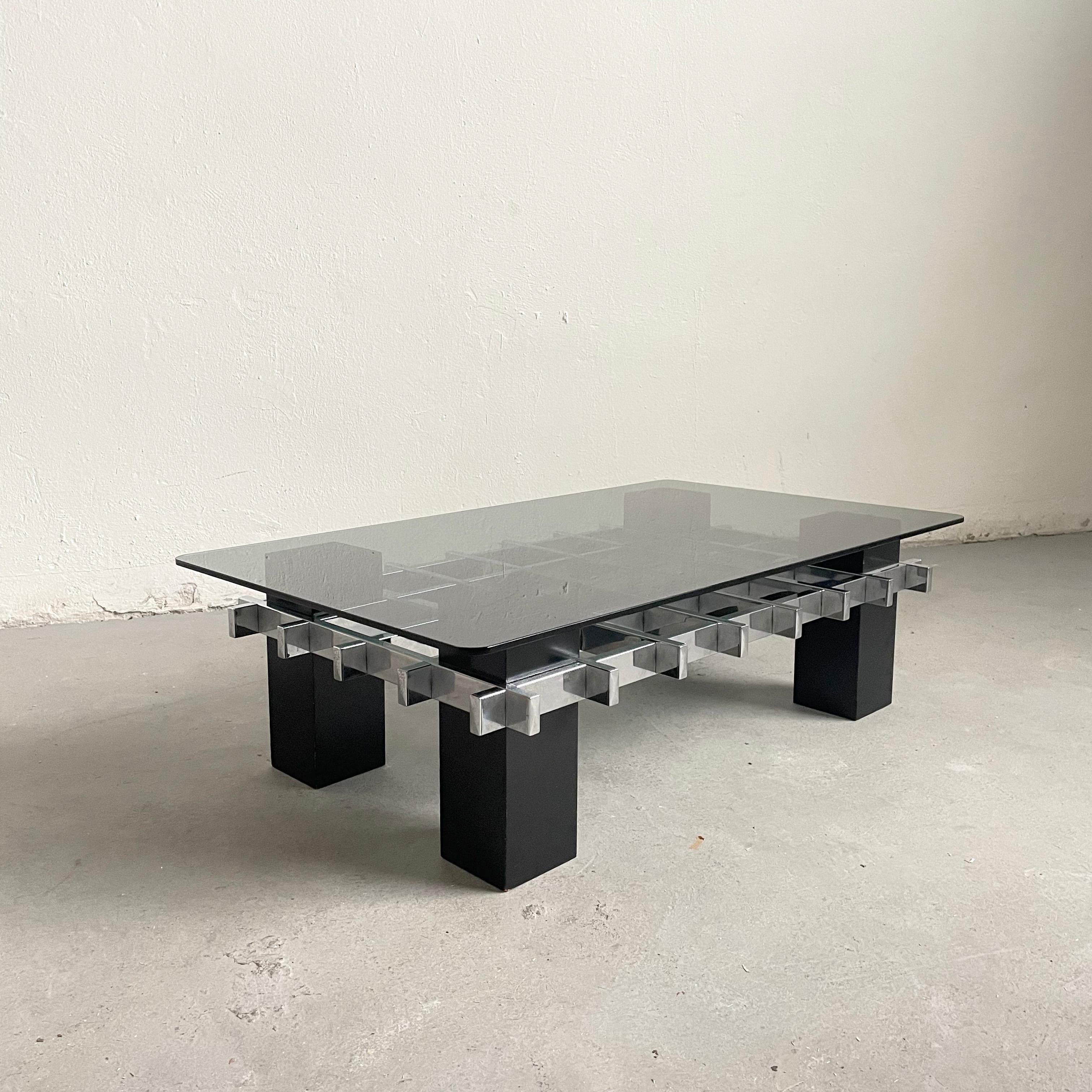 A beautiful coffee table with a unique geometric design. The design is attributed to Milo Baughman and it is made in the USA around the late 1960s or early 1970s. 

It has rectangular black wood legs that support a rectangular tinted glass plate