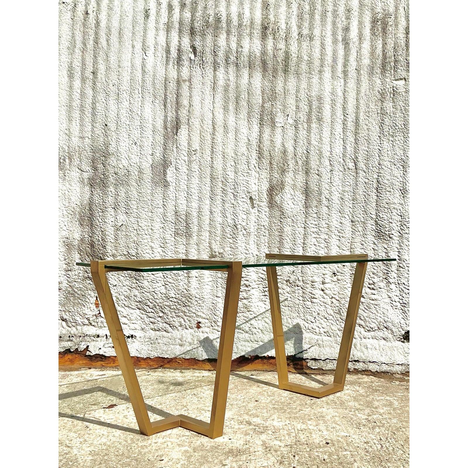 Dieser auffällige, zeitgenössische Konsolentisch im Vintage-Stil vom Design Institute of America (DIA) strahlt mit seinem schlanken, vermessingten Metallsockel und der eleganten Glasplatte moderne Raffinesse aus. Das geometrische Design mit offenem