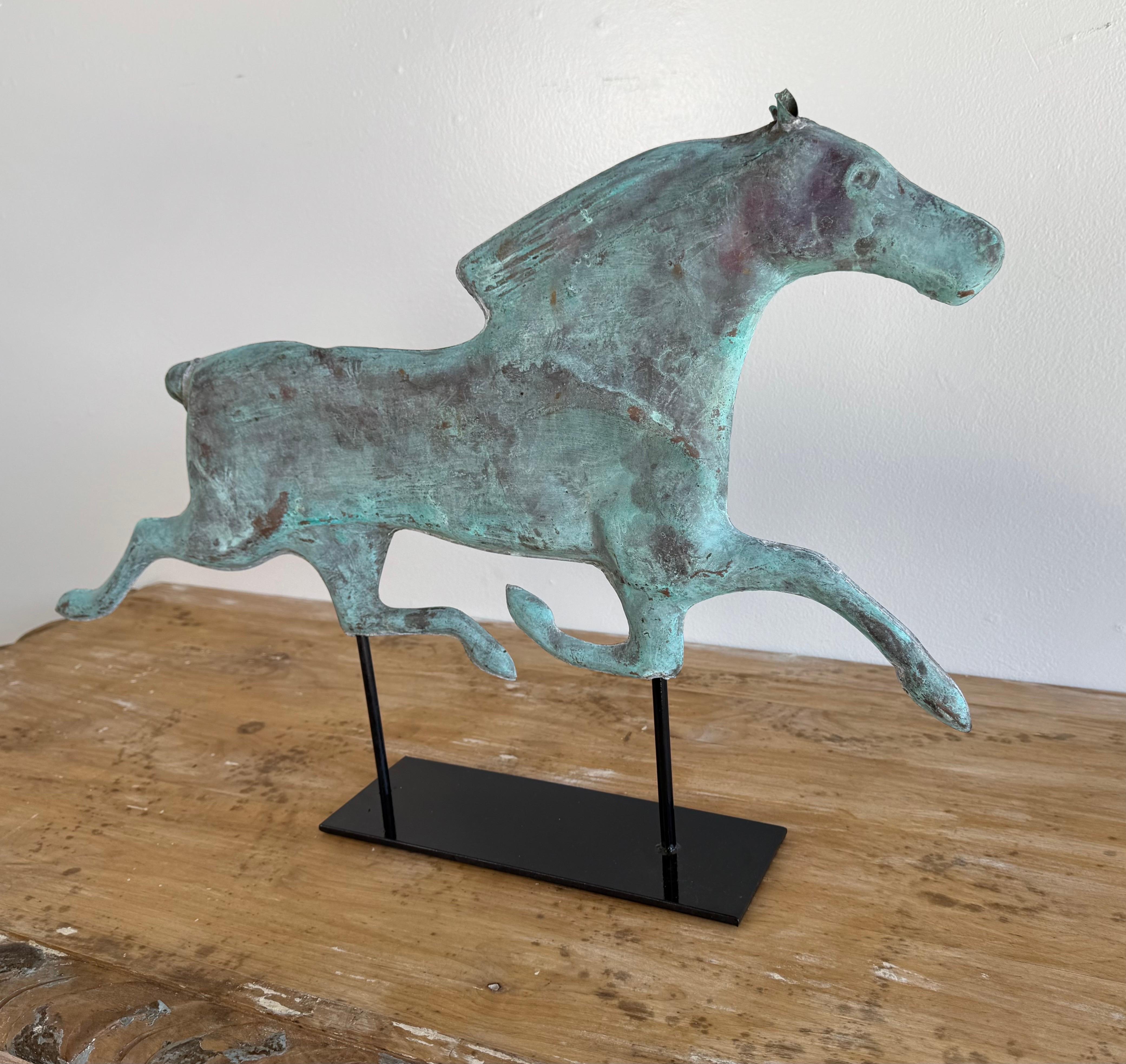 This sculptural piece is a vintage copper horse weather vane, artfully displayed on a modern black metal stand. The figure captures a horse in mid-gallop, with fluid lines that convey movement and energy. The surface features a beautifully aged