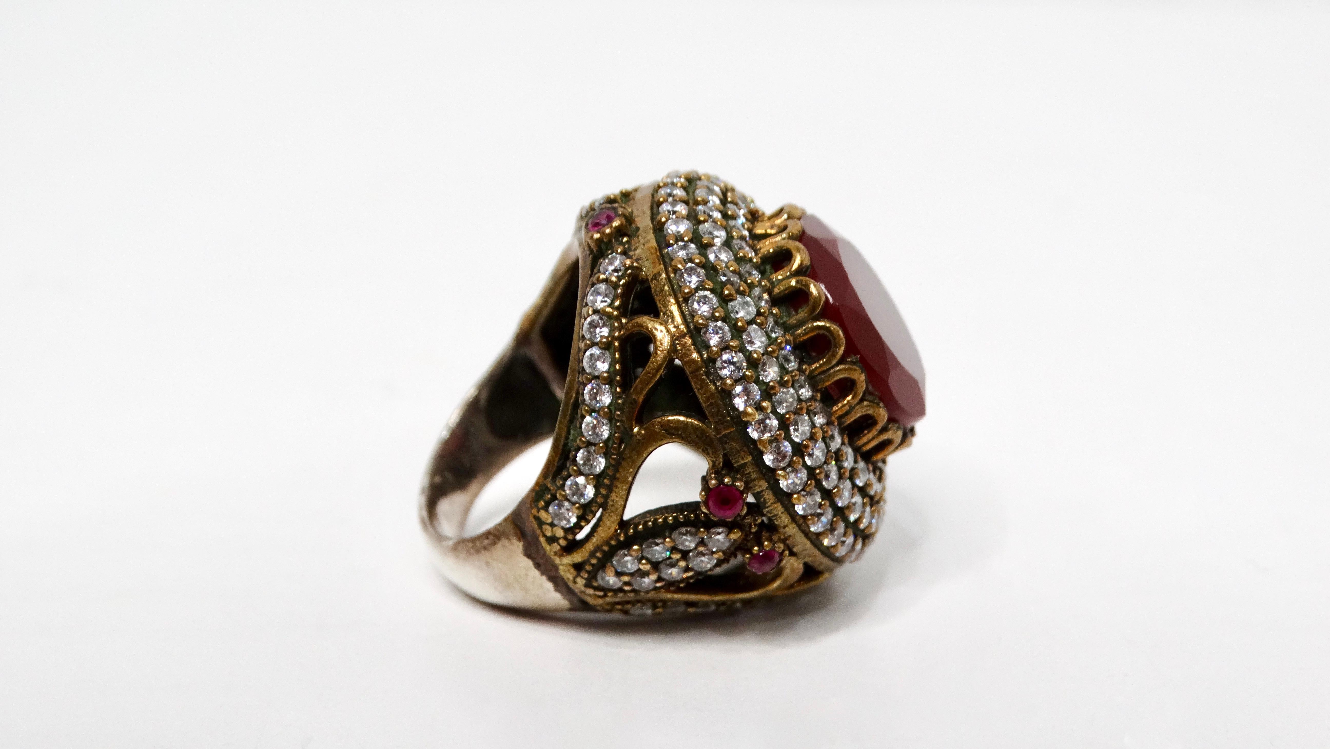 Elevate your jewelry collection with this Vintage Costume Ruby & Rhinestone Detailed Round Statement Cocktail Ring, a stunning blend of elegance and bold design. This ring is a mixture of sterling silver and brass. At its center is a striking round