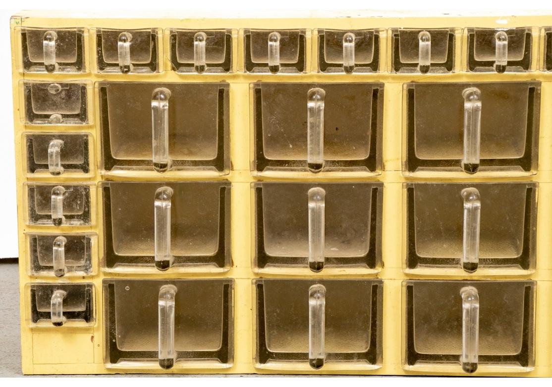 A large and authentic yellow painted pine counter top cabinet housing multiple glass drawers for various small sundries. The top row and along the left side are small drawers, along with three tiers of larger drawers. All glass drawers are textured