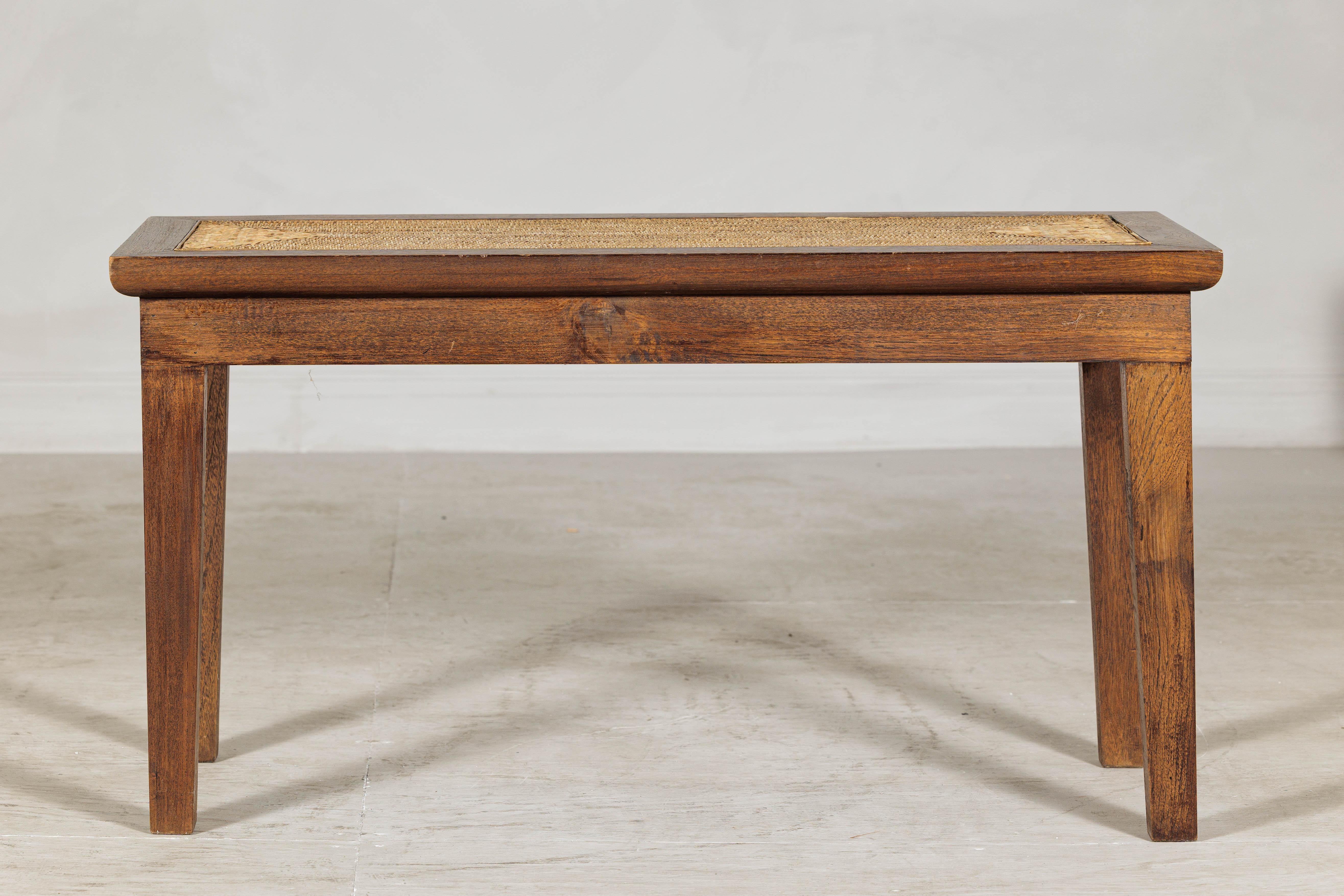 This vintage Asian bench is crafted from solid hardwood with a warm, medium-toned finish and features a handwoven rattan panel inset into the rectangular top. The clean lines and tapered legs speak to a restrained, utilitarian design sensibility,