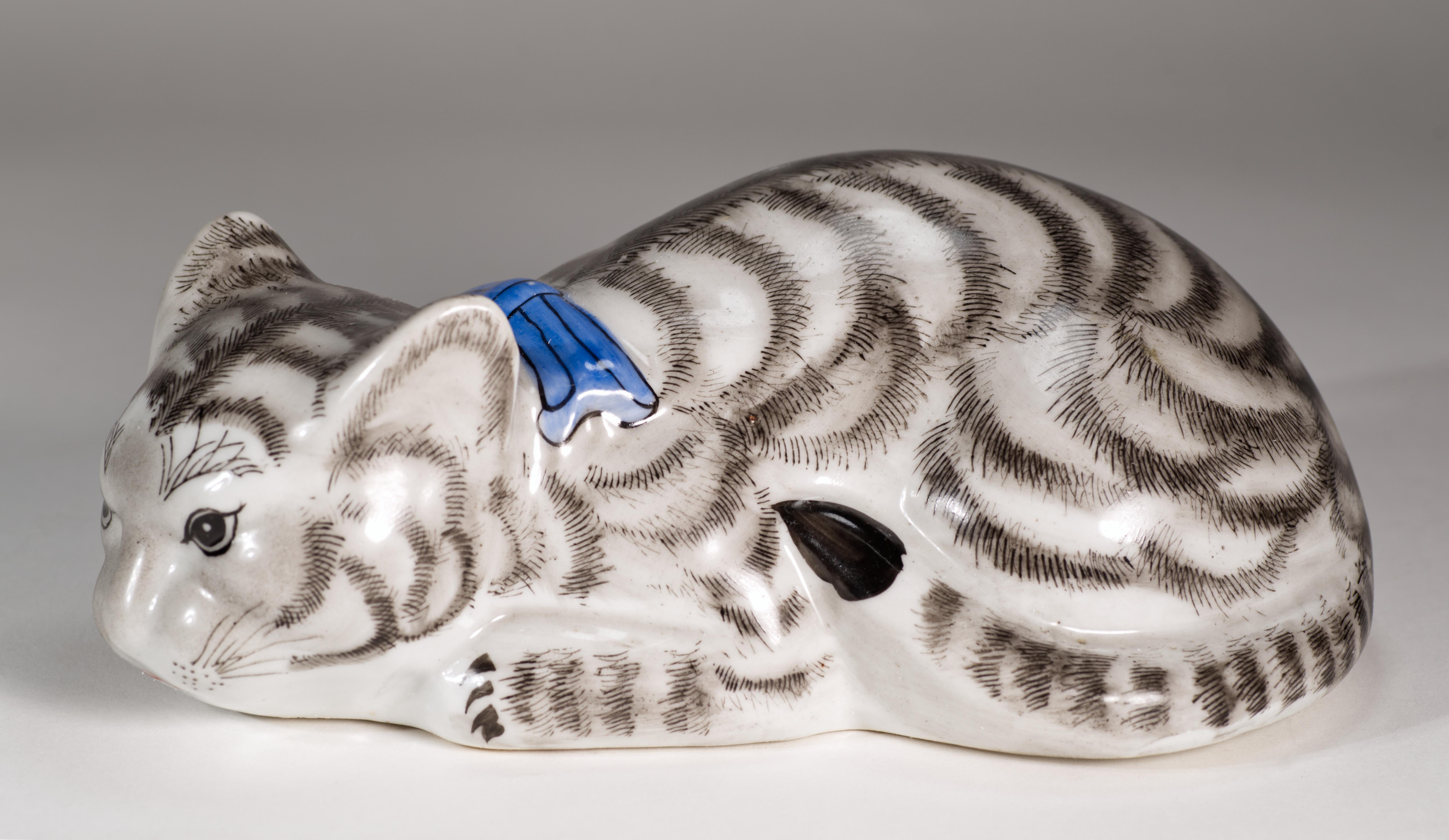 Figurine vintage en porcelaine ou en céramique représentant un chat en position accroupie, en train de chasser et prêt à bondir sur sa proie. La fourrure de chat blanche et gris foncé est rehaussée d'un nœud bleu vif. La fourrure est méticuleusement