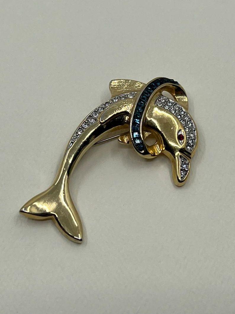 Sencillamente hermoso Precioso broche vintage con forma de delfín. Con un Delfín saltando a través de un Aro, engastado con Cristales Azules. Juego de Manos Delfín con detalles de Cristal Centelleante. Montaje dimensional en tono dorado. Mide aprox.