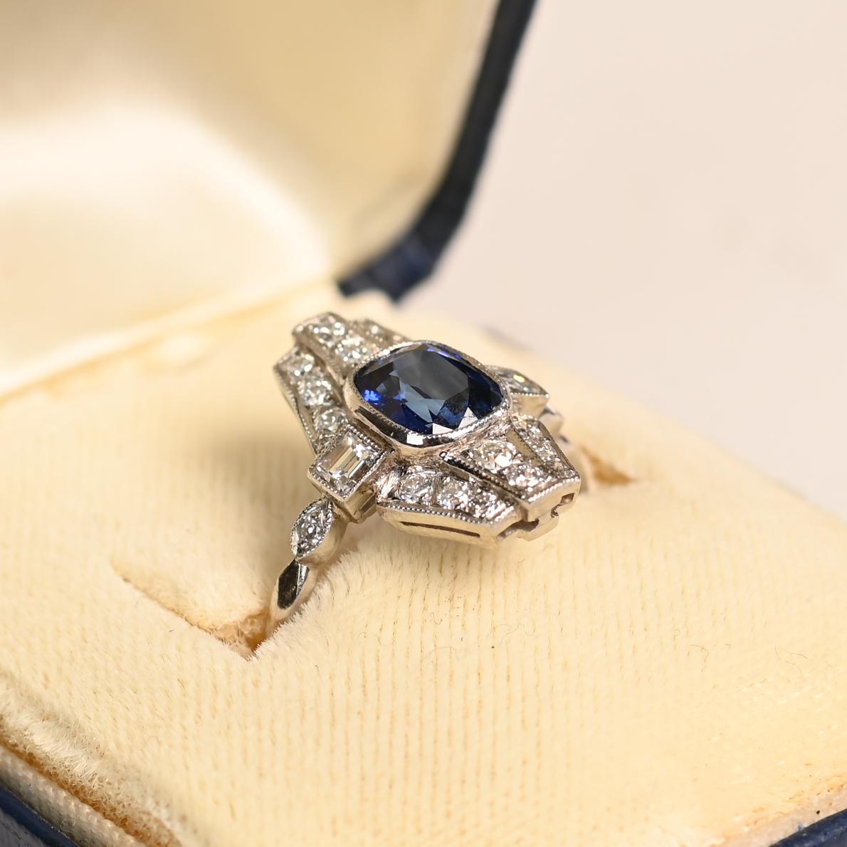 Questo squisito anello allungato mette in mostra un sorprendente zaffiro naturale di circa 1,2 carati con taglio a cuscino, che irradia una saturazione blu profonda e accattivante. Lo zaffiro è completato da una splendida aureola di diamanti antichi