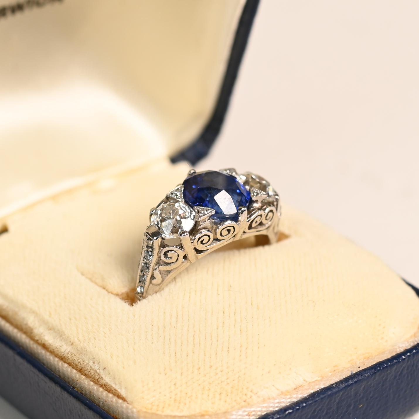 Questo squisito anello a tre pietre mette in mostra un raro zaffiro di Ceylon da 3,06 carati con taglio a cuscino, incastonato nel platino con delicate griffe triangolari ornate da diamanti a taglio singolo. Lo zaffiro blu è affiancato da due