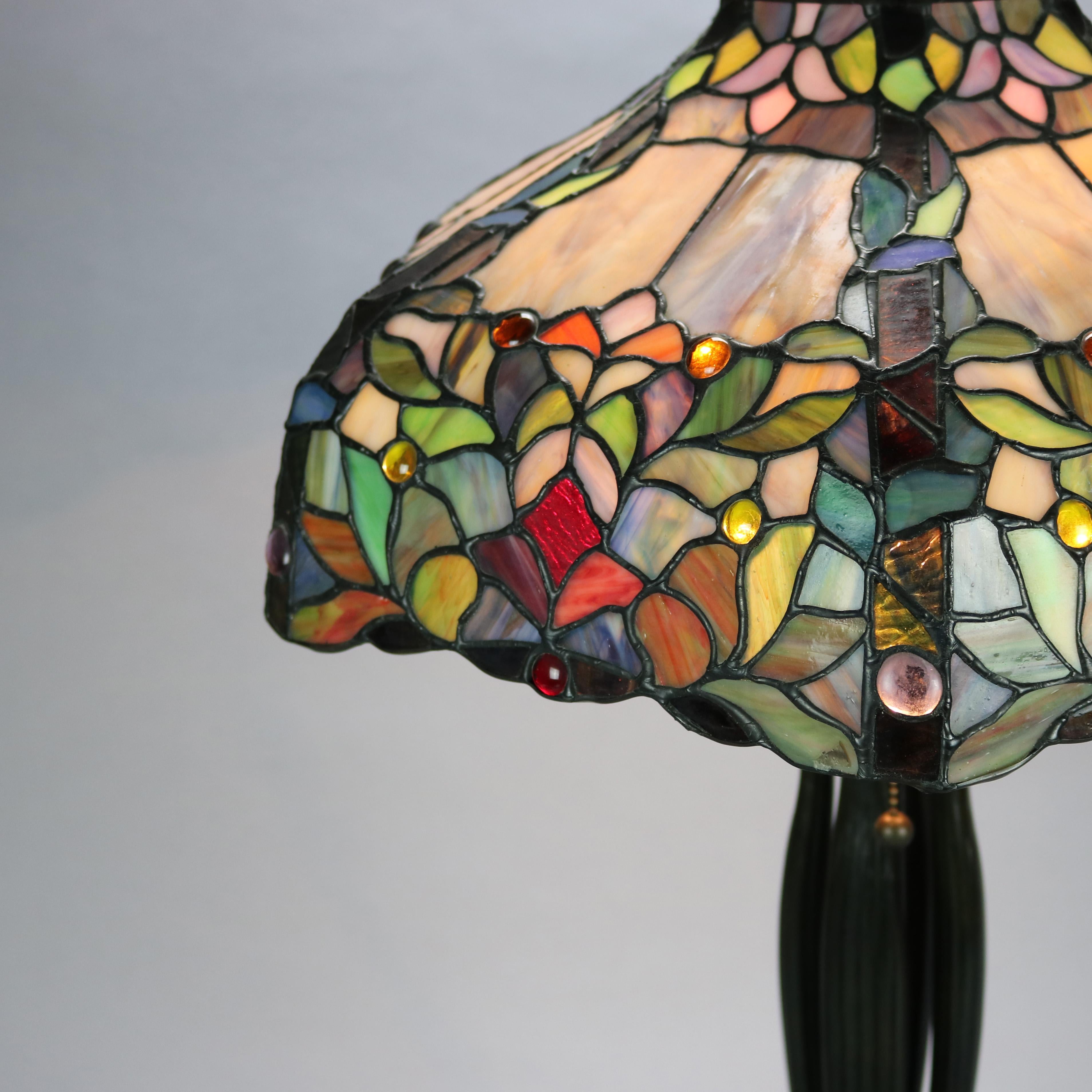 An vintage table lamp by Dale Tiffany offers an Art Nouveau design with stylized parasol form shade in mosaic leaded stained and jeweled glass foliate design over bronzed cast two socket base having column with stylized paw feet, 20th