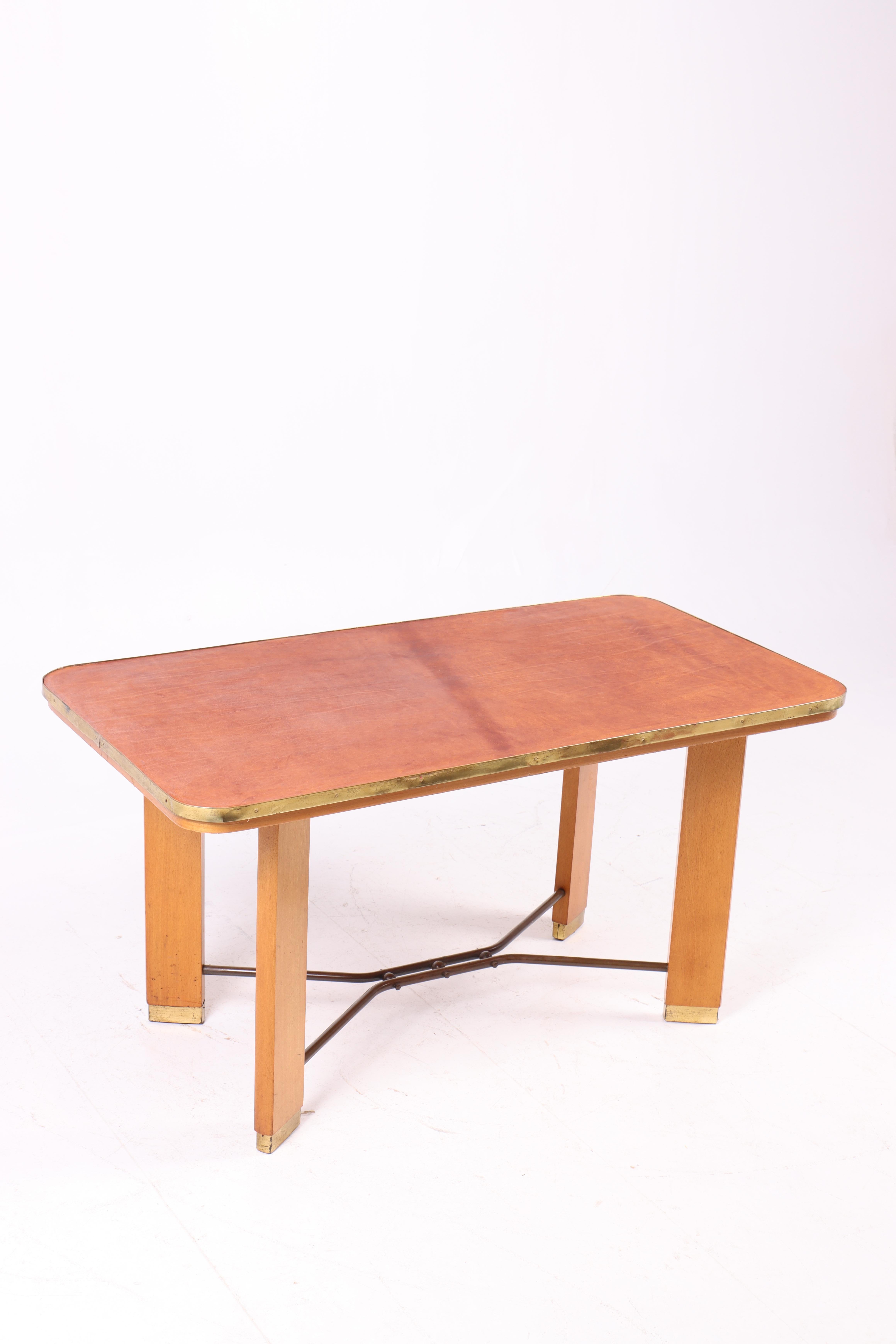 Esta mesa de centro de los años 50 está fabricada en madera de haya con un bonito tablero de cuero patinado, que le da un distintivo aspecto vintage. Además, la mesa está adornada con elegantes detalles de latón a lo largo de los bordes, que
