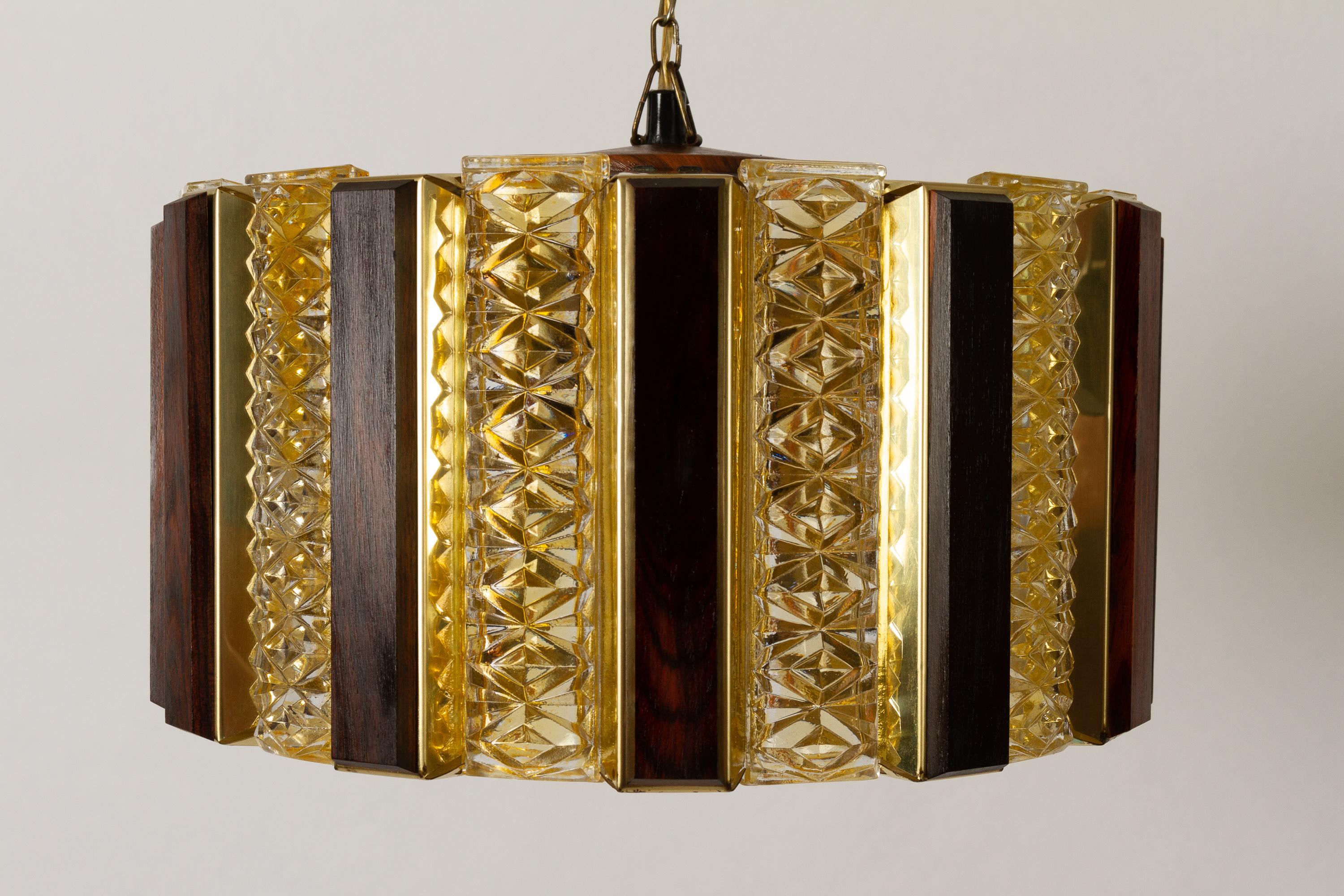Vintage Danish ceiling pendant by Werner Schou for Coronell Elektro, 1960s.
Scandinavian Modern ceiling lamp in brass, glass and teak. Round with alternating yellow/amber colored glass bricks and wooden slats mounted on brass plates. Lamp is