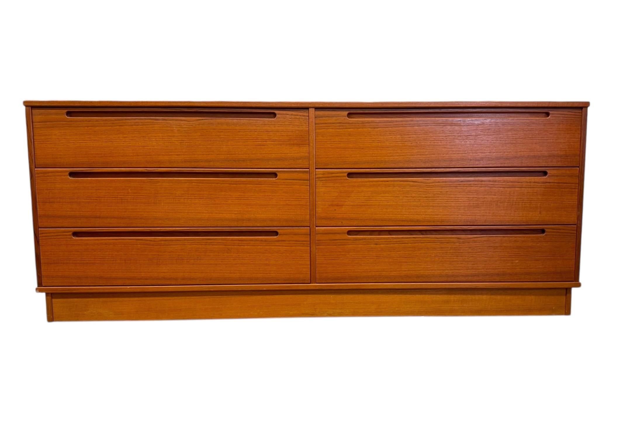 The Vintage Danish Mid-Century Modern Teak Dresser features a sleek, minimalist design typical of the mid-20th century Scandinavian style. Crafted from teak wood, it showcases a warm, rich grain finish with a smooth, polished surface. The dresser