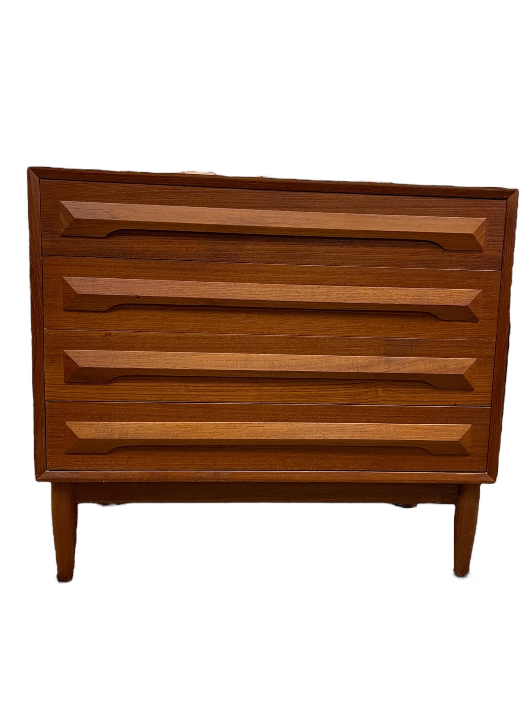 Un comò di medie dimensioni in teak di metà secolo con maniglie scolpite e importato dalla Danimarca. Quattro ampi cassetti a coda di rondine, con gambe affusolate. Usura coerente con l'età e l'uso. Piccoli segni e lievi graffi come da foto e video.