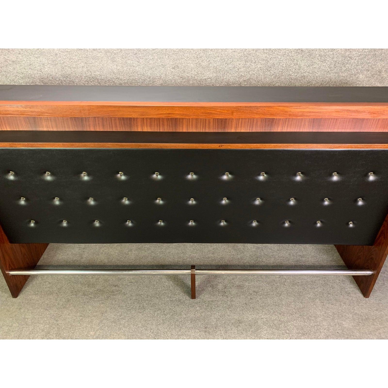 Here is an exceptional Scandinavian Modern bar and 4 stools manufactured in Denmark in the 1960s.
This large bar features a vibrant rosewood grain, a tufted front in black Naugahyde, a chromed footrest at its bottom and many large cocktails
