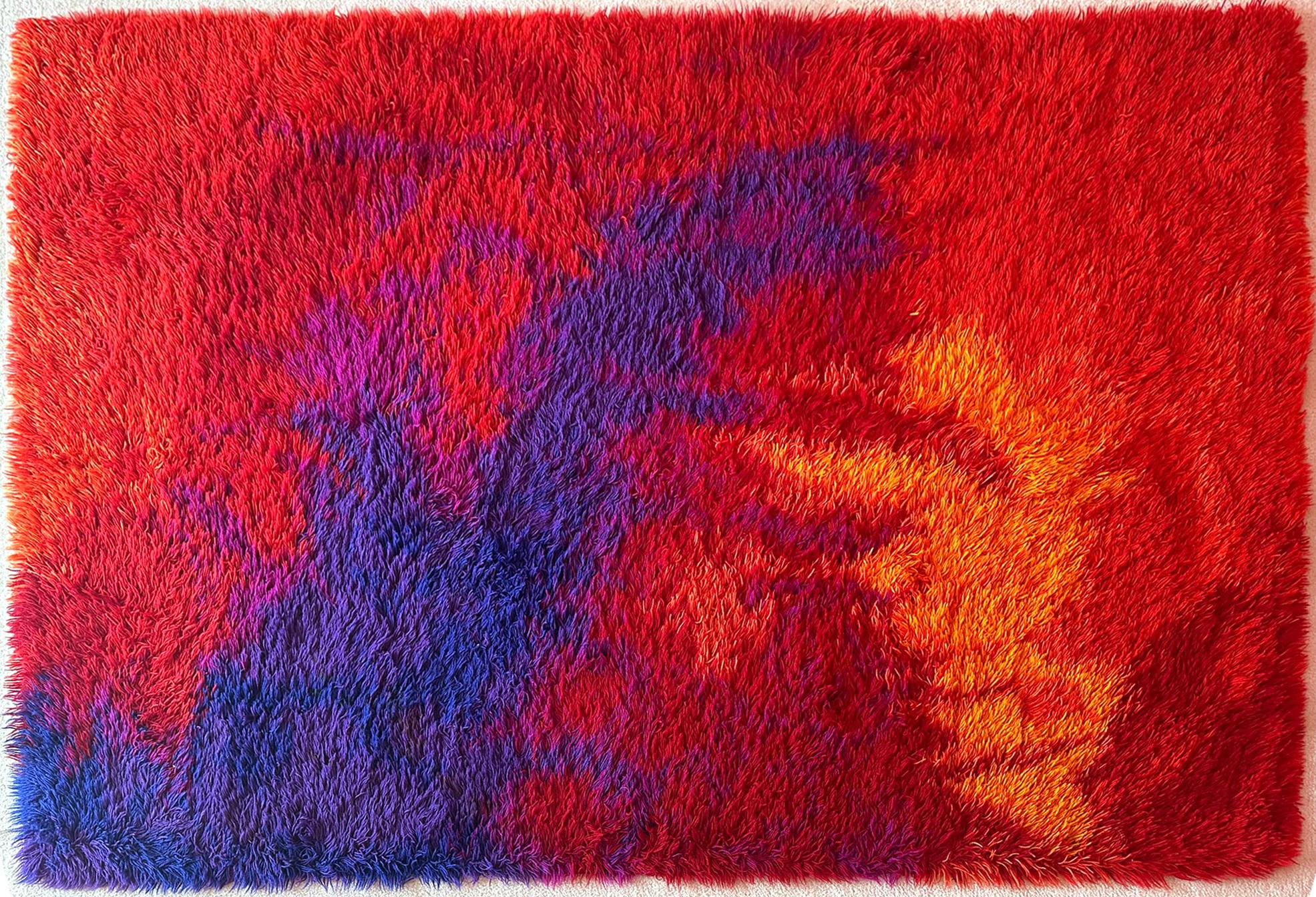 A vintage Danish area wool rug made by Unikataeppe in Denmark circa 1960s. The bright colored rug features long shaggy fibers and remains in very good condition. On the background of mottled red colors in subtle different shades, splashes of cobalt