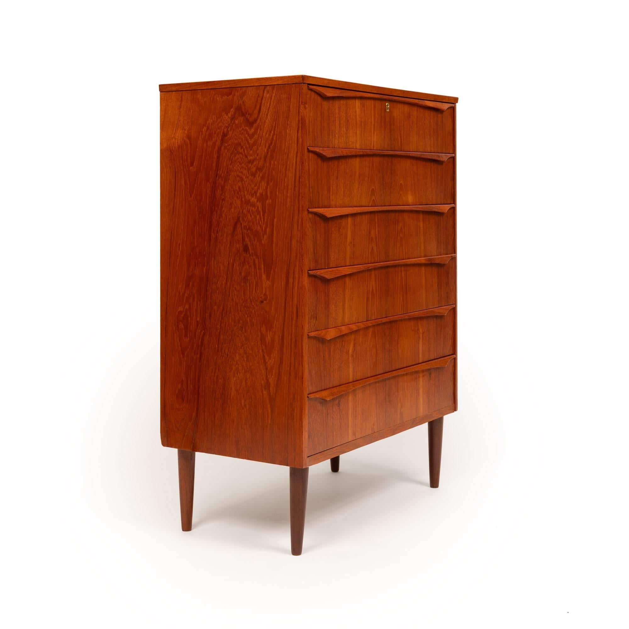 Esta impresionante cómoda tallboy de teca danesa de mediados de siglo de 1960 es una pieza de artesanía atemporal. Con exquisitas vetas de madera y tiradores de teca tallados a mano, esta cómoda alta ofrece seis cajones espaciosos para elevar tanto