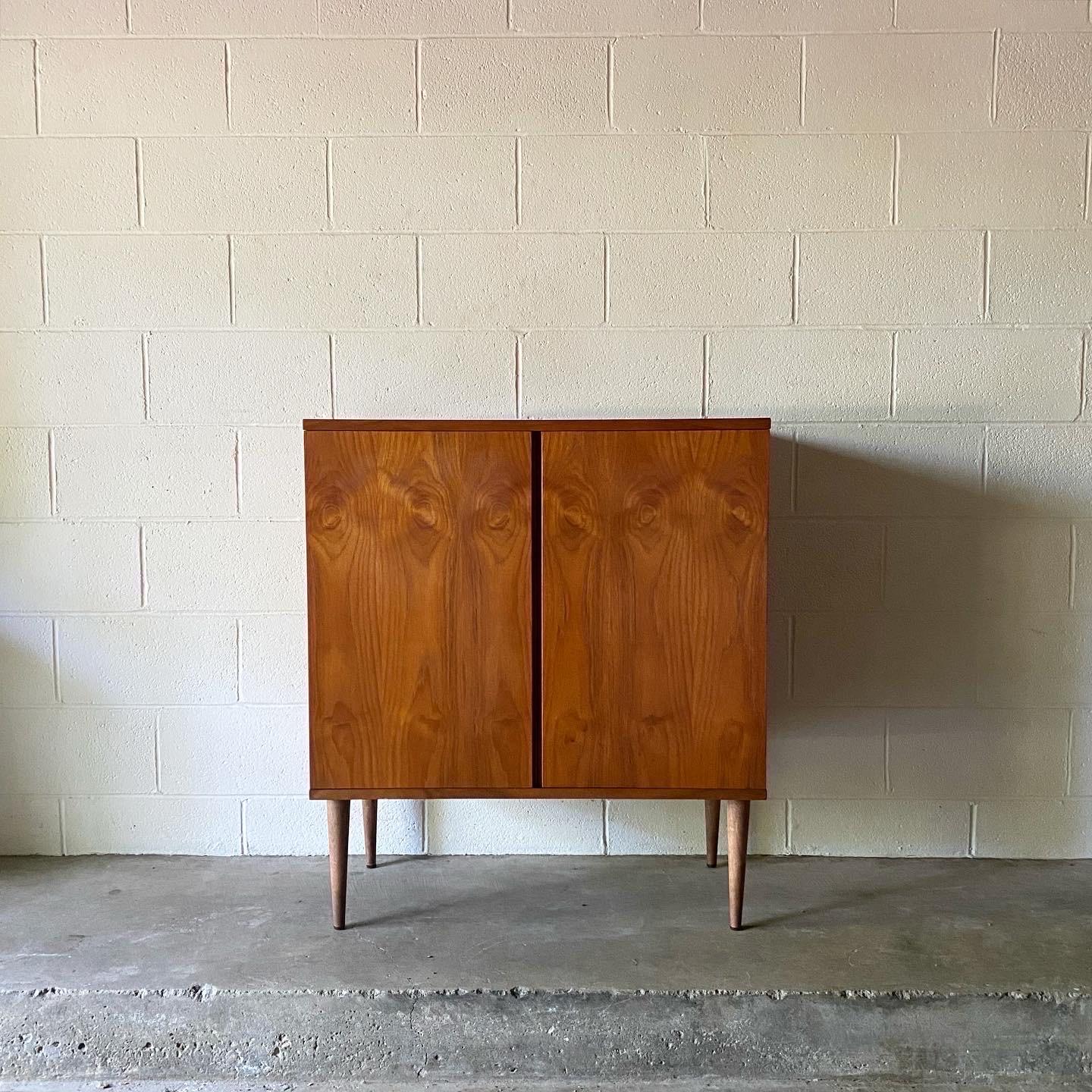 This is a great teak sideboard or bar cabinet designed by Hans Olsen, made in Denmark ca. 1960’s. This beautifully crafted piece displays opposing teak veneers and brass fittings. Doors swing open to reveal an adjustable shelf. Perfect for an