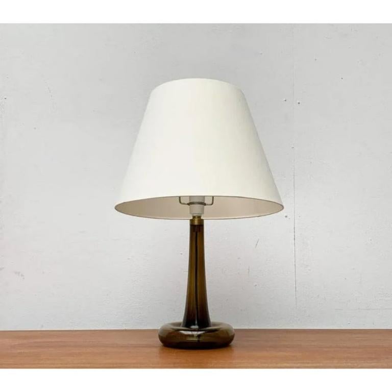 Élégante lampe de table du fabricant danois Holmegaard d'après un design de Michael Bang des années 1970.

Un projet dans un langage formel traditionnel. Cette lampe à poser convainc par son pied élégant en verre coloré. Le parapluie simple présente