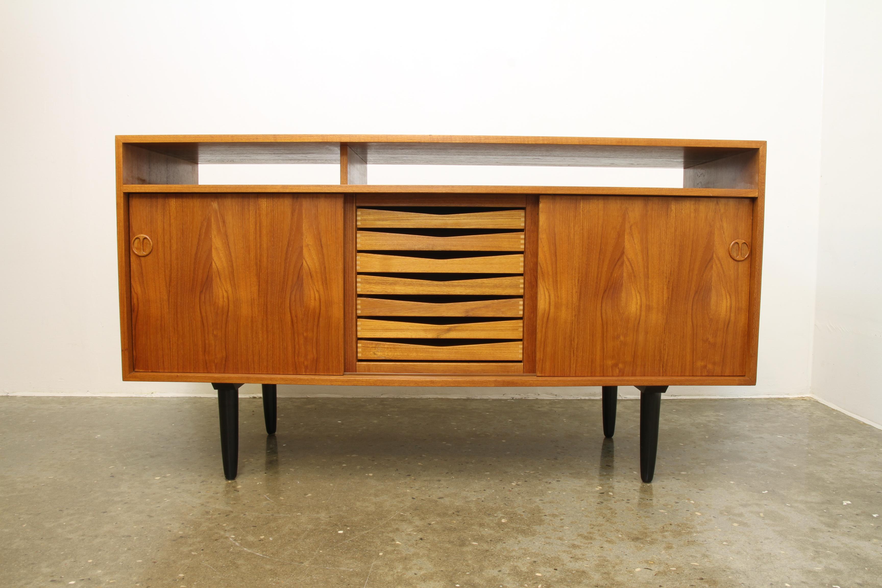 This low stylish Danish midcentury credenza in teak features two compartments with shelves, and a drawer unit in the middle. The drawers has beautiful visible joints. Loads of storage space. The open shelve is perfect for HiFi equipment.
Backside