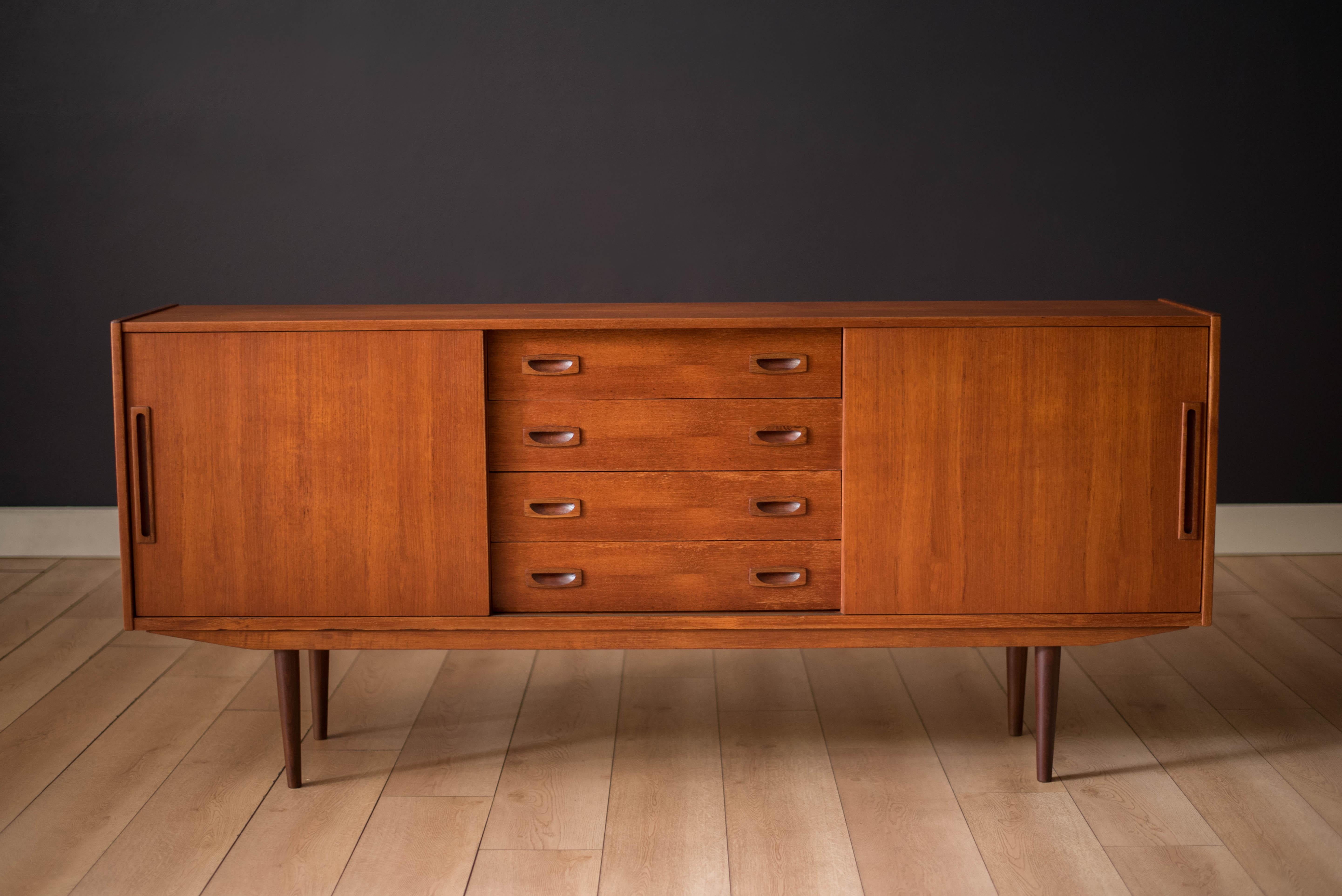 Mid-Century Modern sideboard credenza in teak circa 1960's. This showcase storage piece features four dovetailed drawers accented with solid teak afromasia sculpted handles. Sliding doors reveal two interior cabinets with a removable single shelf.