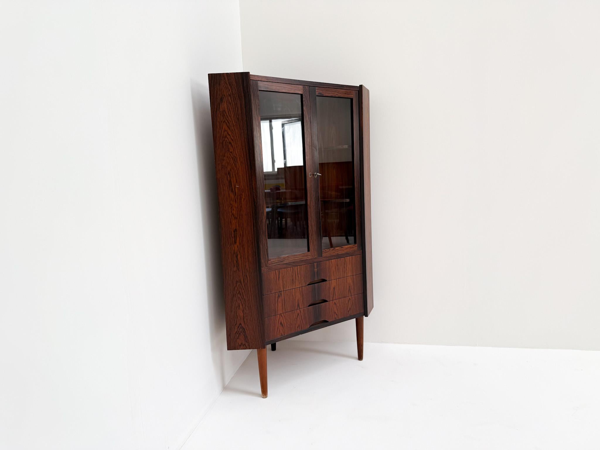 Striking vintage corner cabinet crafted in highly figured rosewood veneer, made in Denmark in the 1960s. This elegant piece features two glass doors, three lower drawers with signature sculpted handles, and beautifully tapered solid wood legs. The