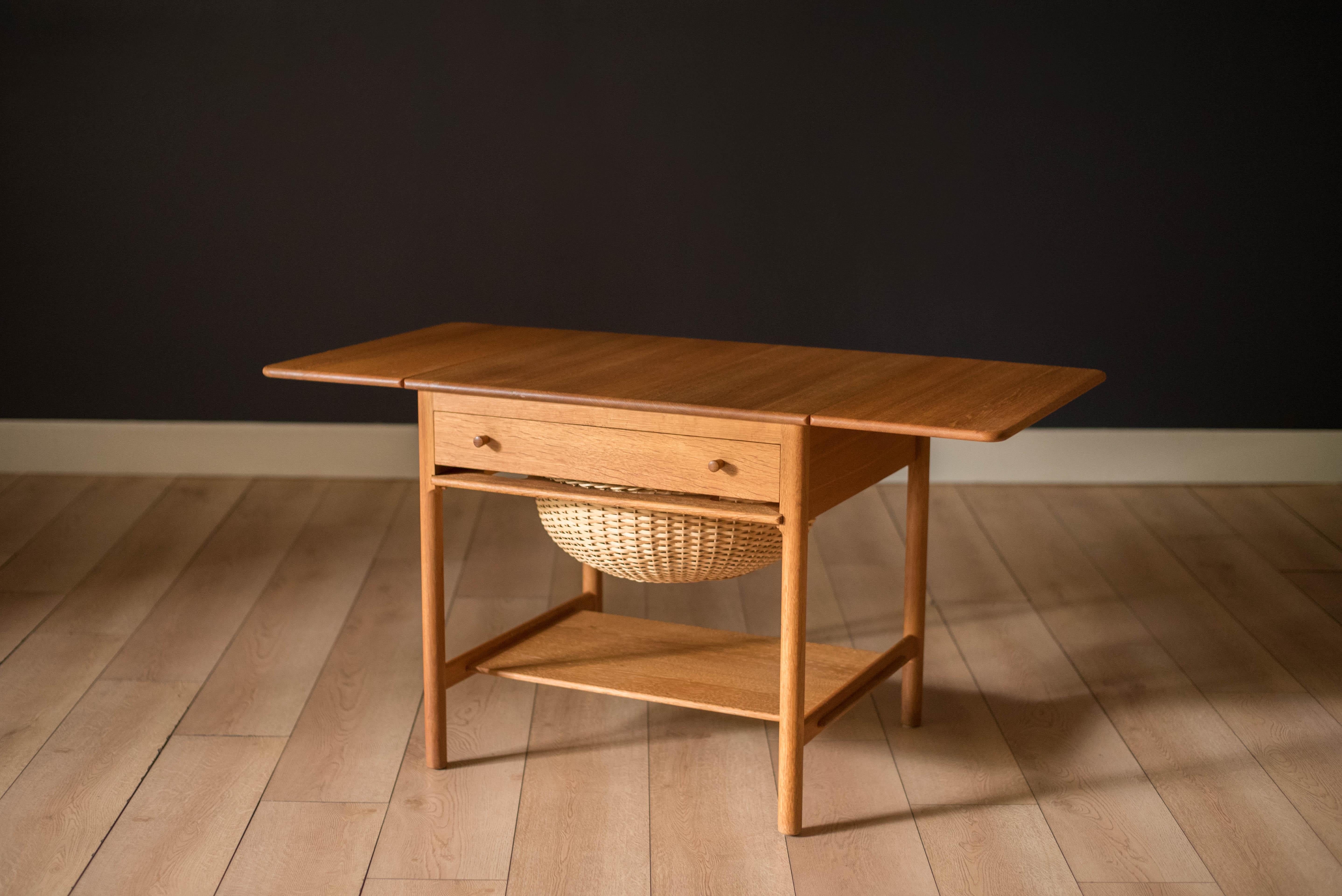 Mid Century Danish modern sewing table designed by Hans Wegner for Andreas Tuck circa 1950's, Denmark. This unique piece is constructed of solid oak featuring an extendable drop leaf tabletop. Includes one dovetailed drawer, a pull-out wicker basket