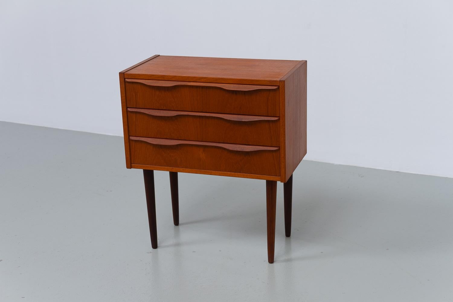 Vintage Danish Teak Bedside Table, 1960s.

Small Scandinavian Mid-Century Modern three drawer dresser in teak veneer. Suitable as a nightstand or side table. Made in Denmark in the 1960s.
Tall round tapered legs in solid teak. Sculpted drawer pulls