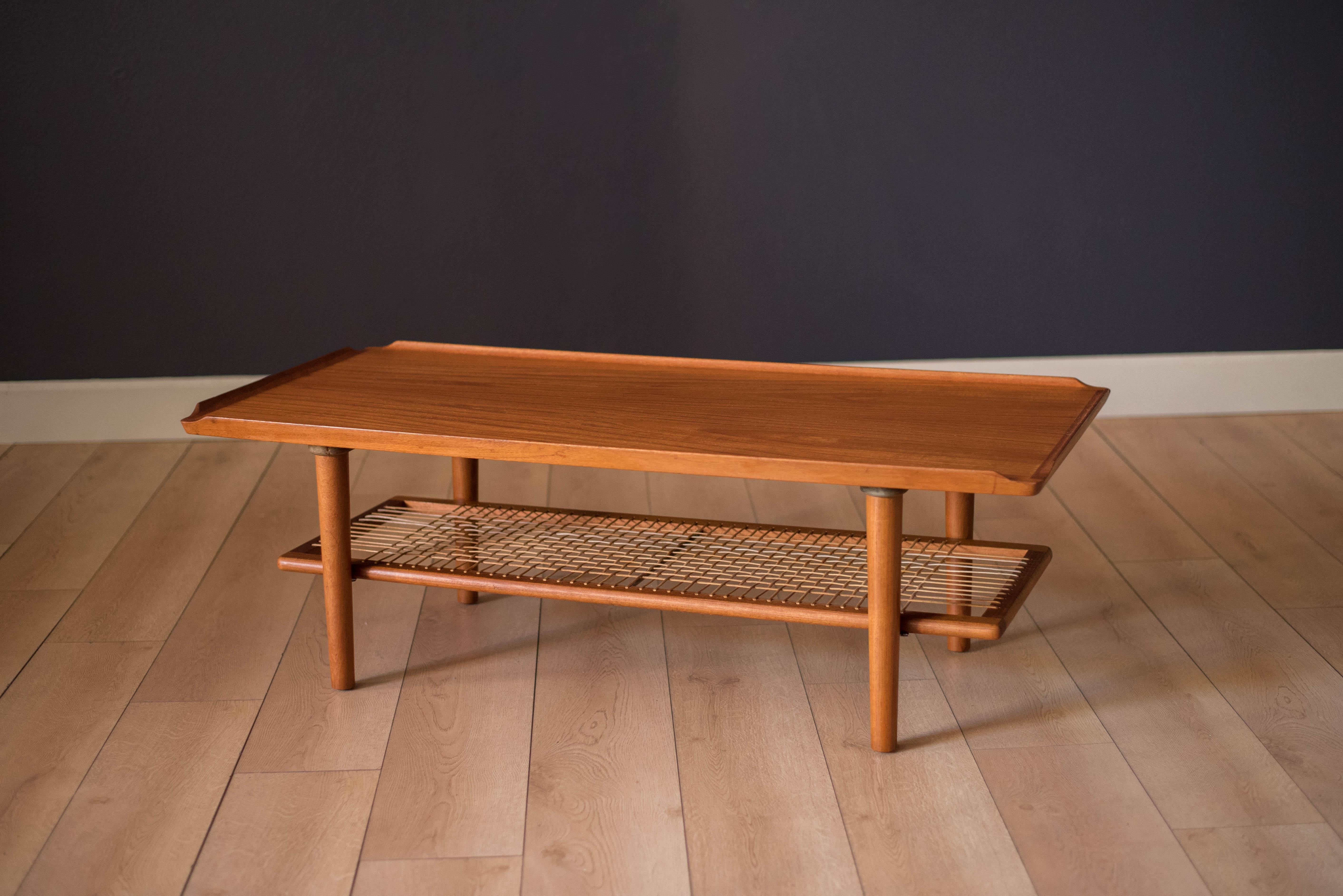 Mid-Century Modern coffee table attributed to Georg Jensen for Kubus. This piece features a unique sculpted border and a lower woven cane magazine shelf. Displays well from all angles.