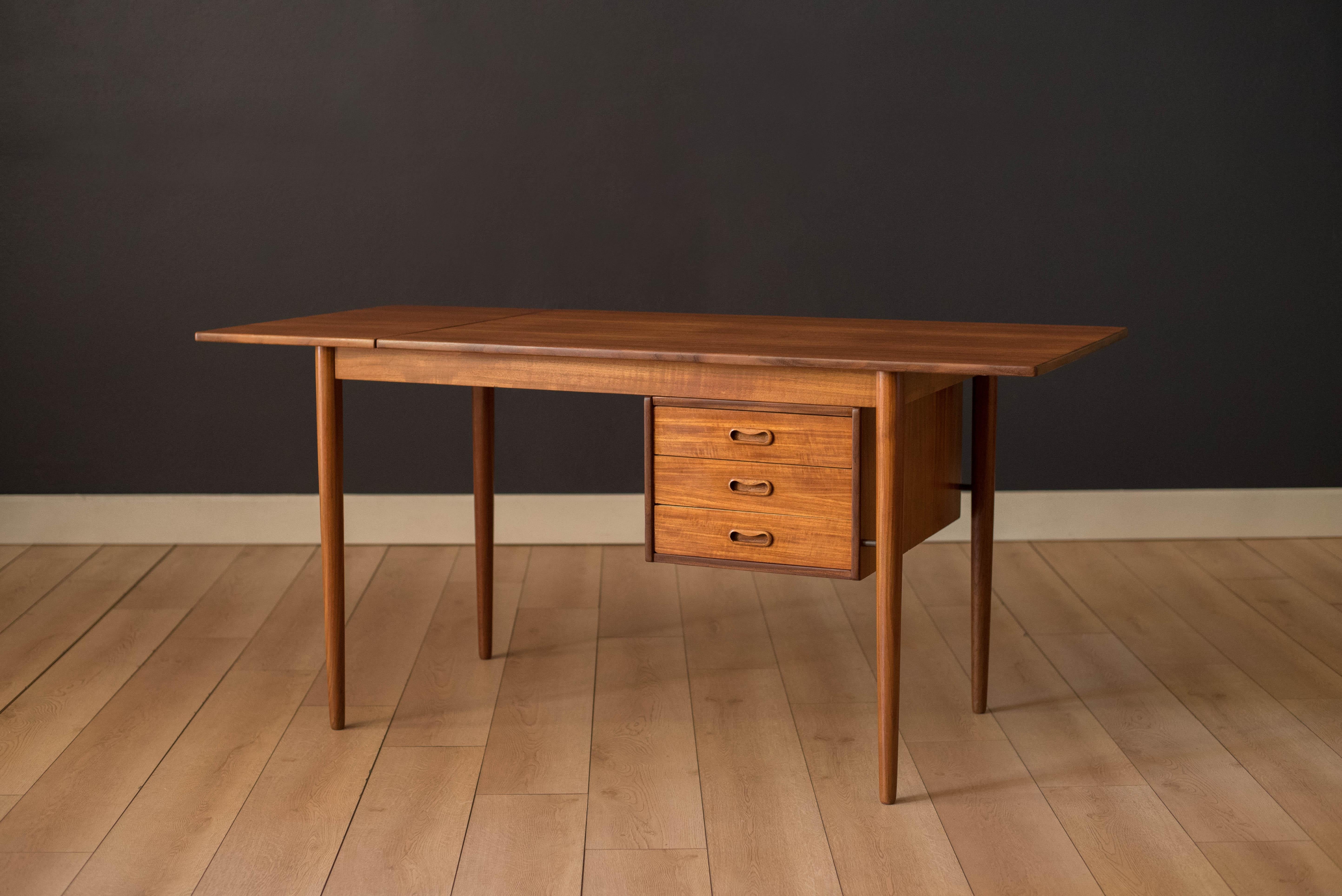 Mid-Century Modern drop leaf desk in teak, circa 1960s. This versatile piece includes three dovetailed with sculpted wood handles. Worktop area expands in size by sliding the drop leaf extension. Accented with brass hardware that attaches to the