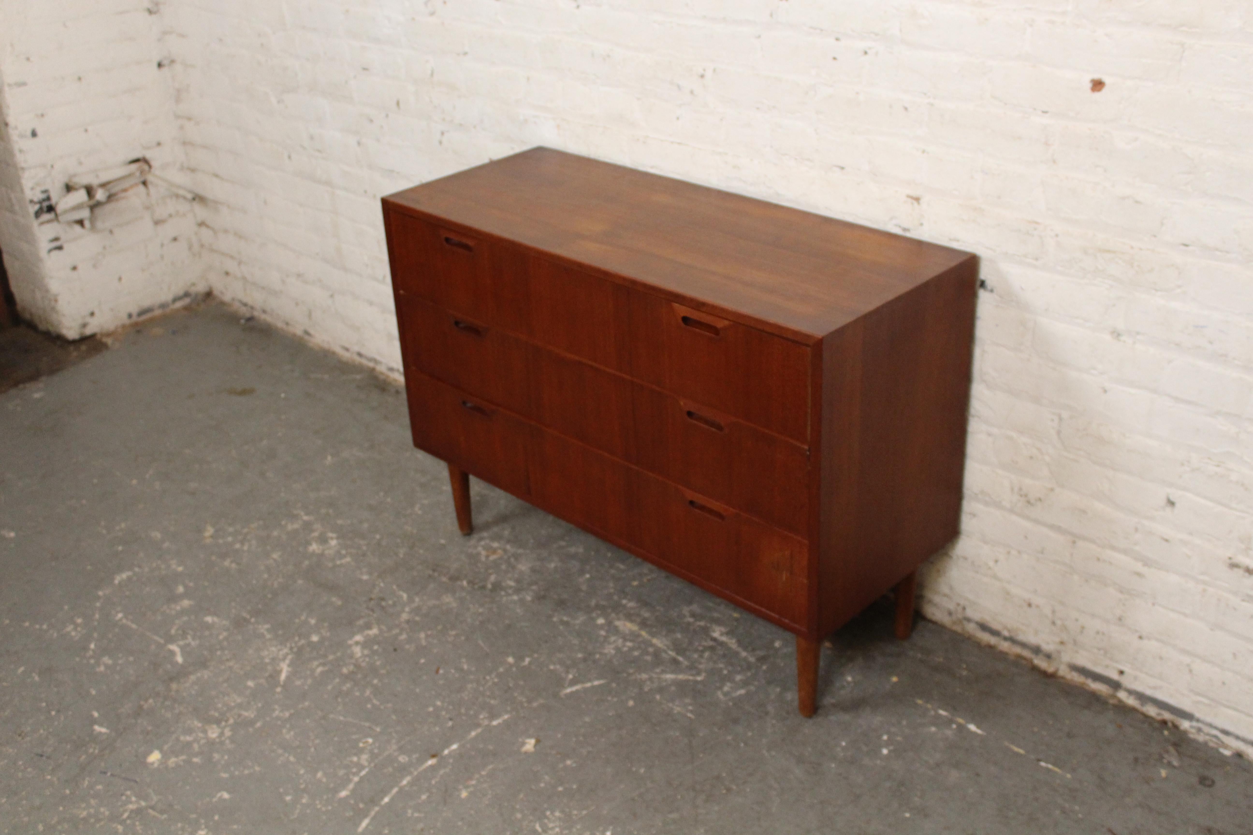 This petite dresser designed by Sven Ellekaer for Albert Hansen and sold by Raymor features three wide drawers with wood handles, teak wood grain throughout and tapered cone legs. A versatile design with removable feet and integrated brackets for